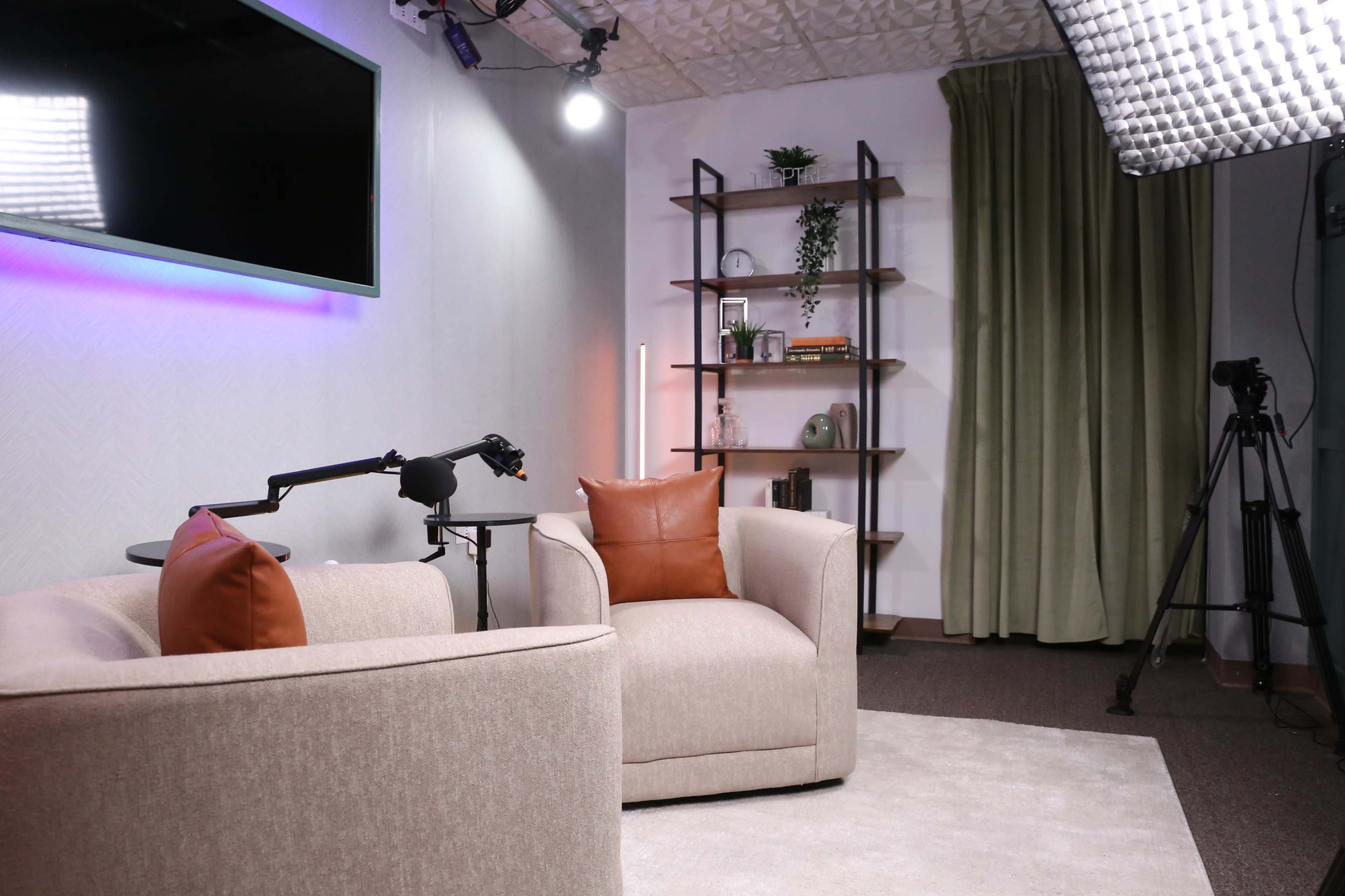 The image shows a well-lit studio setup with two armchairs, a microphone on a stand, a shelf with decorative items, and a camera on a tripod in the background.