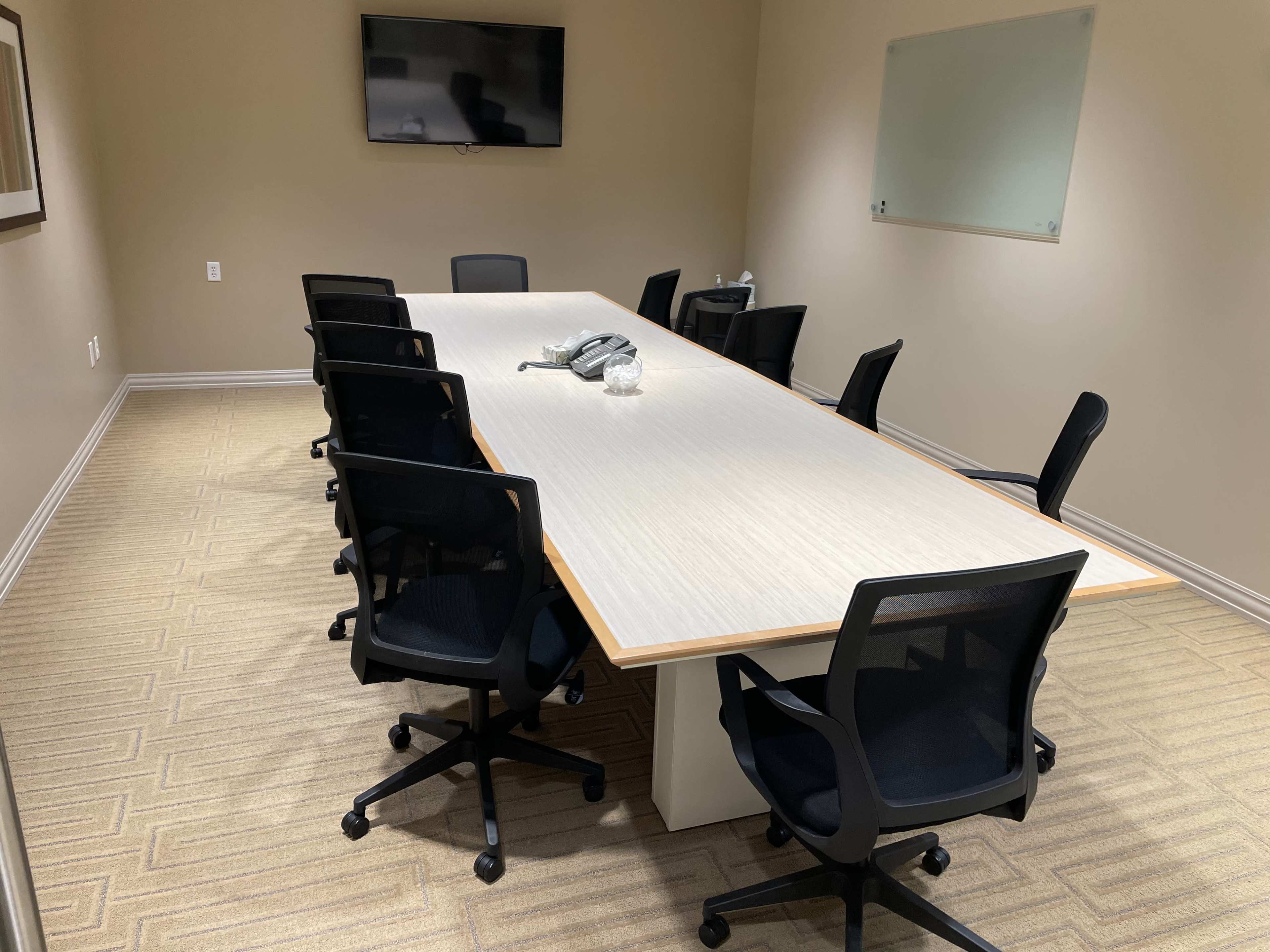 A conference room features a long table surrounded by eight black rolling chairs, with a television mounted on the wall and a whiteboard nearby.