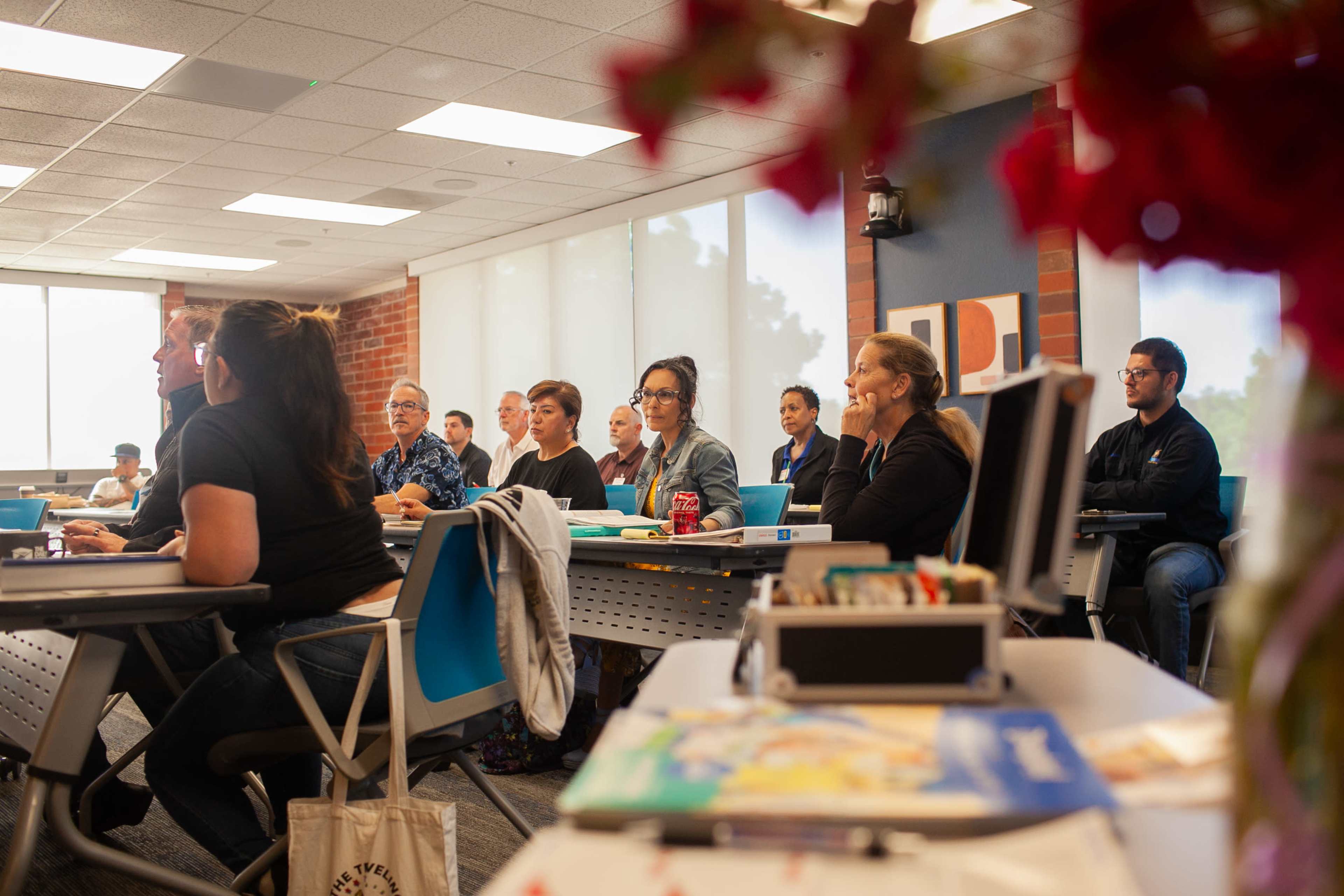A group of students attentively listens in a classroom setting, with a flower arrangement in the foreground.