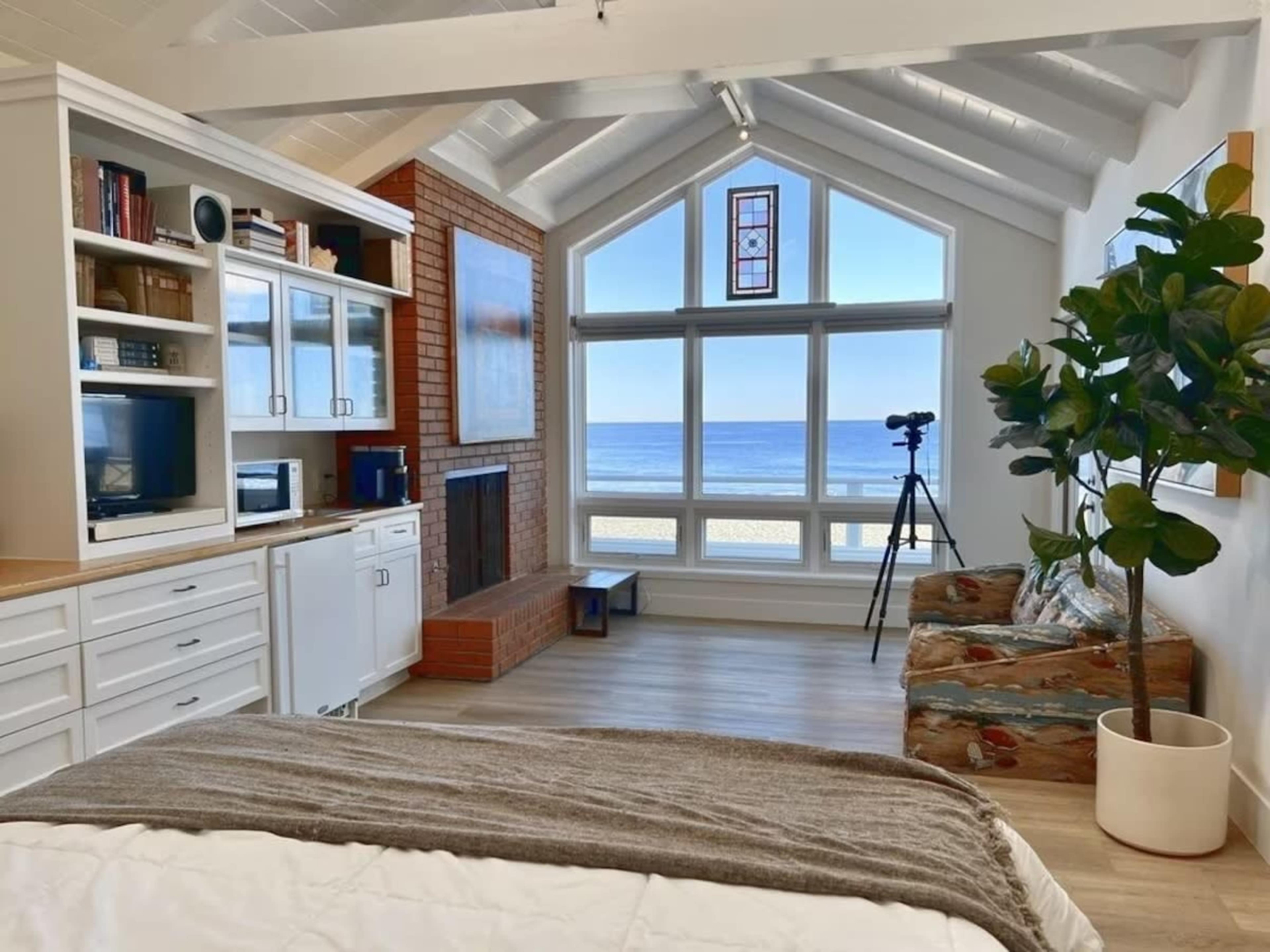The image shows a bright and airy bedroom with a view of the ocean, featuring a bed, a fireplace, a seating area, and large windows letting in natural light.