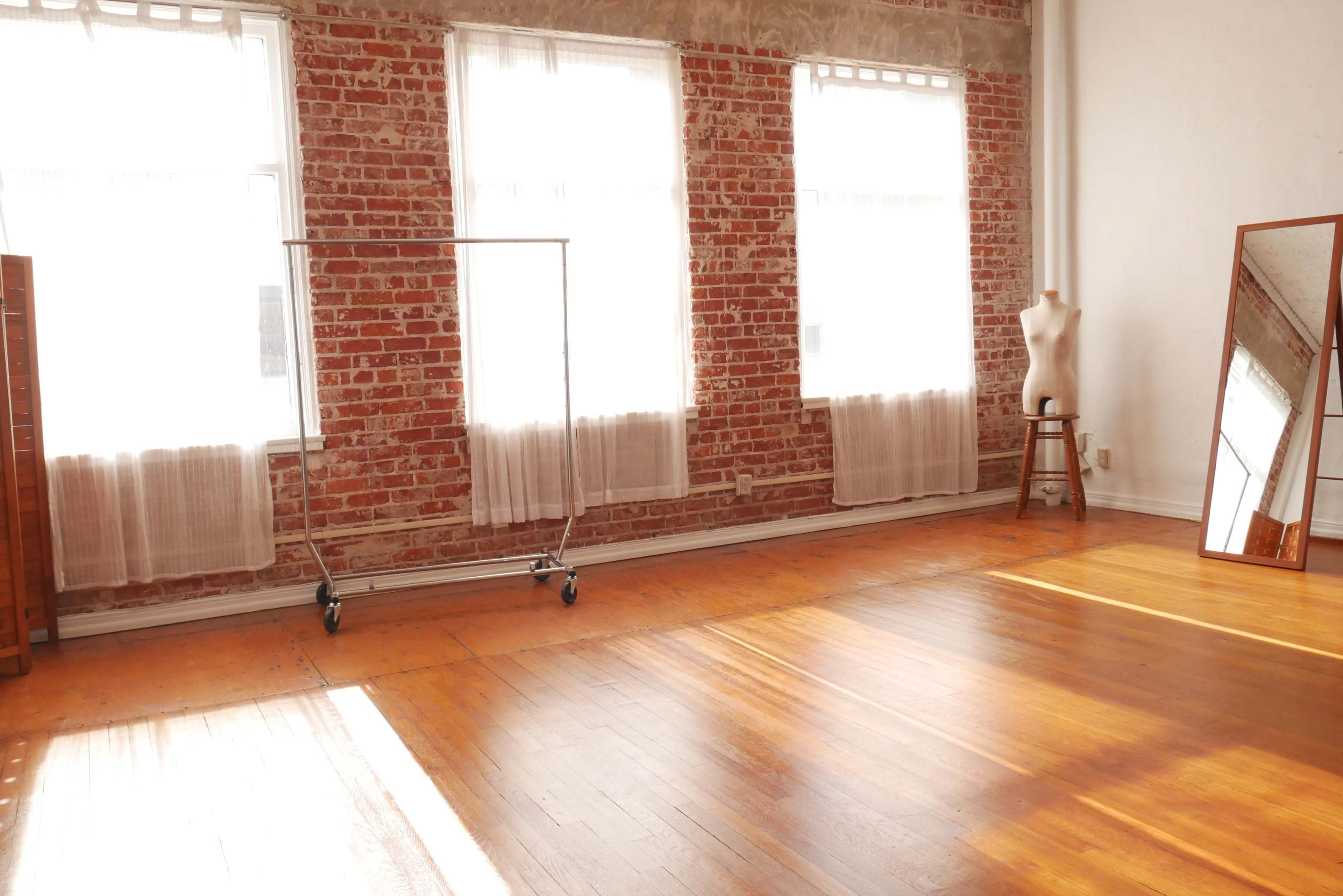 An empty studio space with exposed brick walls, large windows, a clothing rack on wheels, a mannequin, and a mirror reflecting the wooden floor.