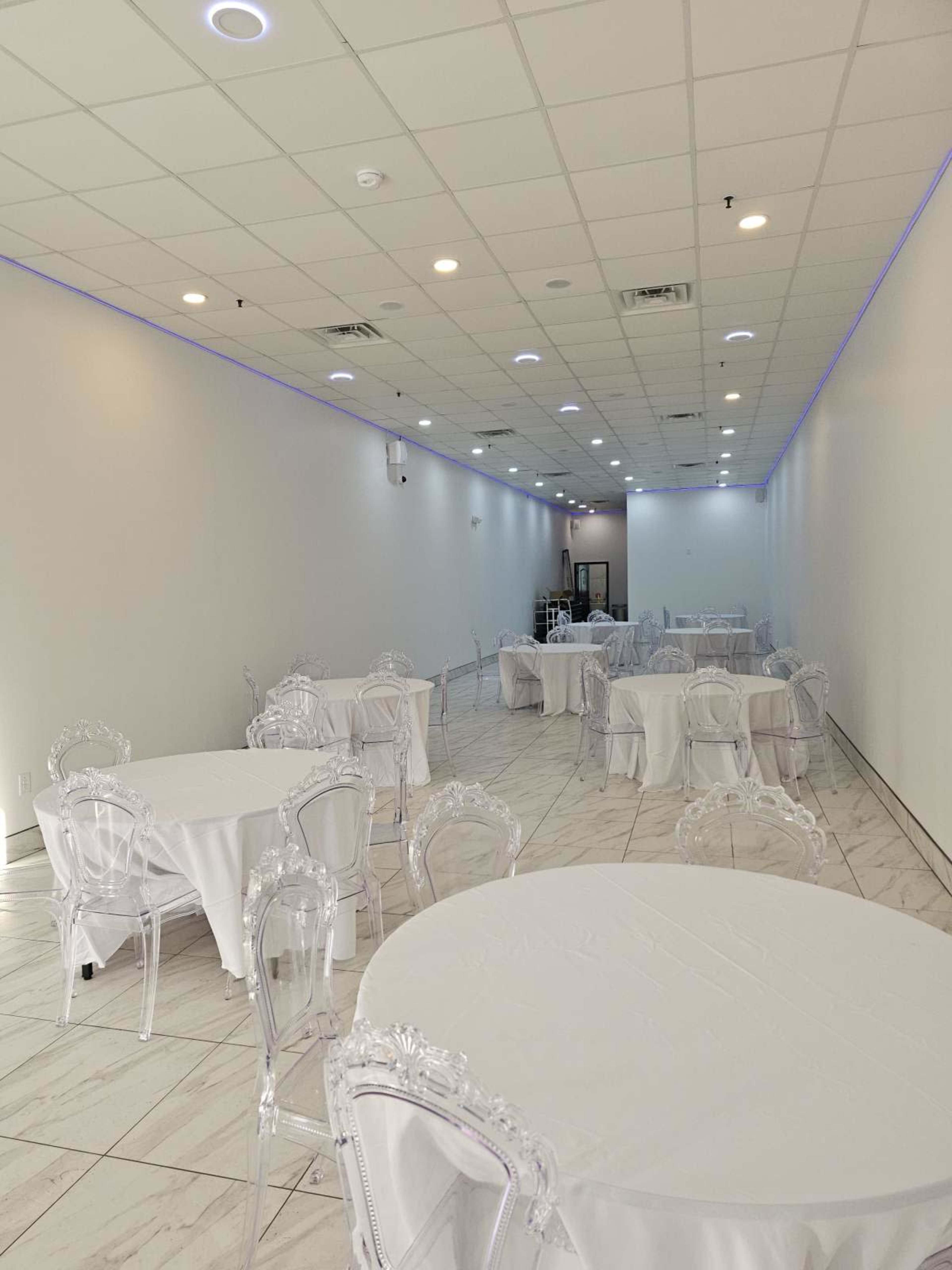 The image shows a large, empty room with white walls and marble flooring, featuring several round tables covered with white tablecloths and transparent chairs.