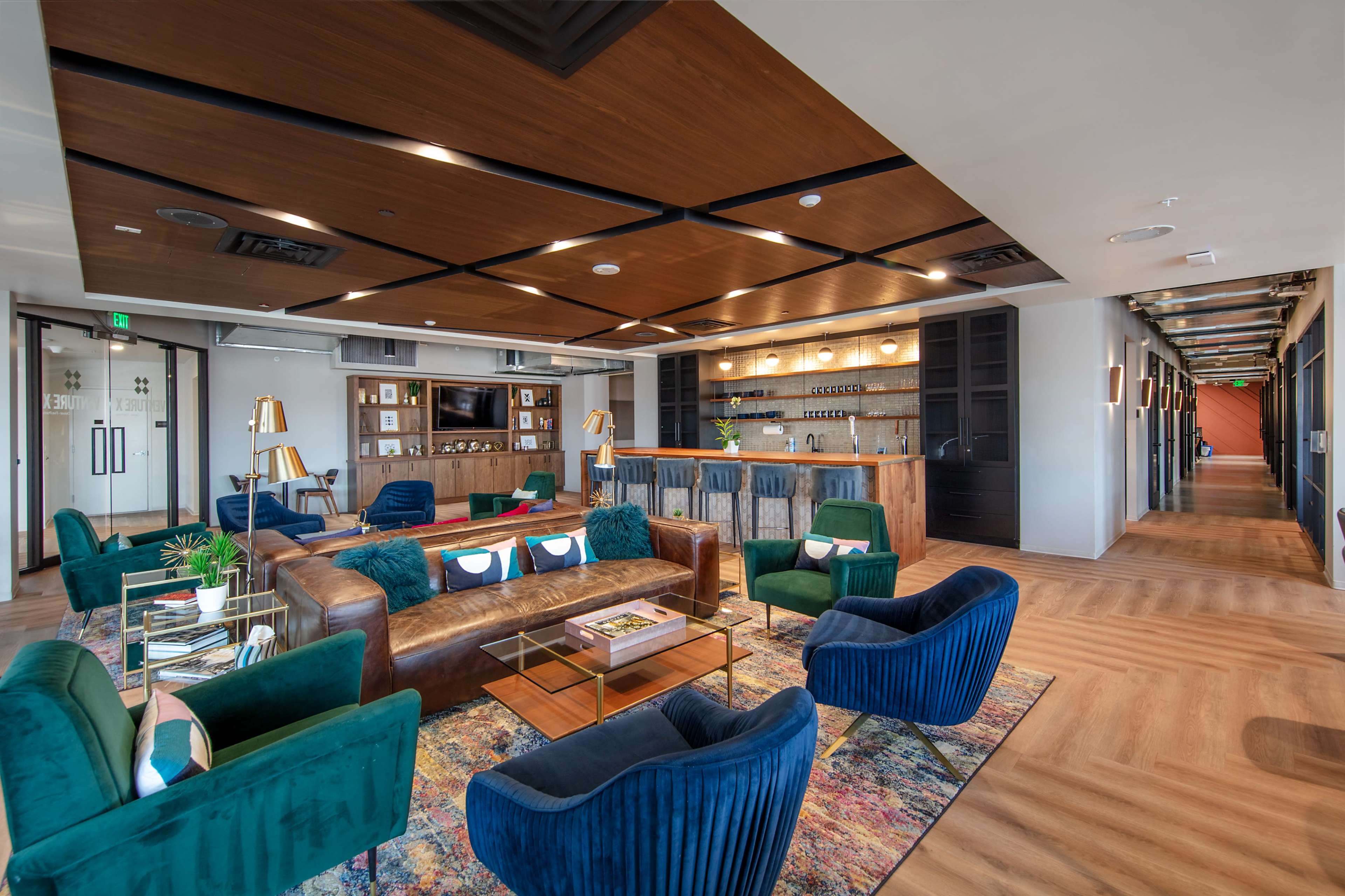 The image shows a modern lounge area with a large sectional sofa, various chairs, and a bar counter against a backdrop of wooden panels and vibrant decor.