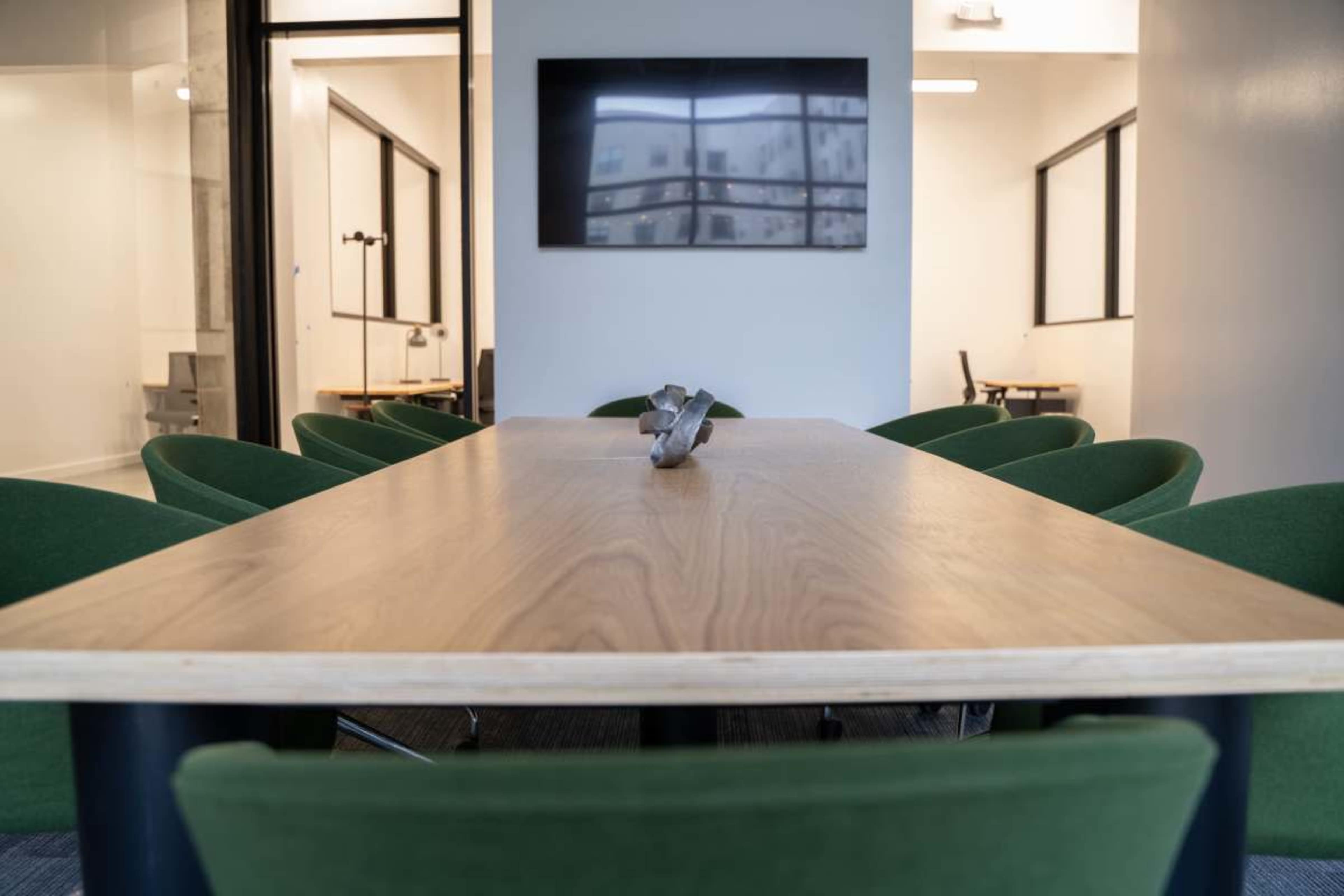 A long wooden conference table is set in a meeting room with green chairs and a large screen mounted on the wall.