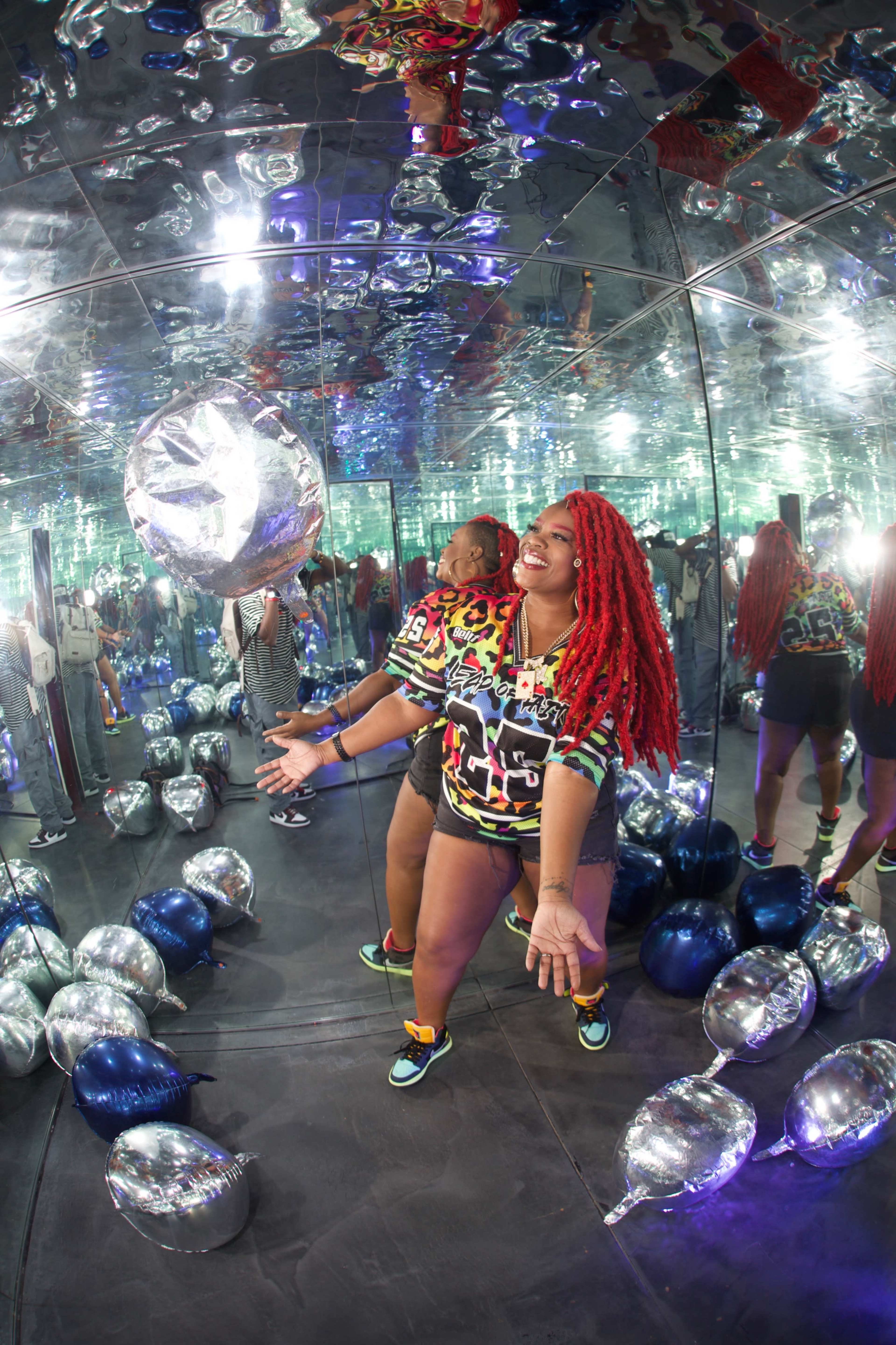 A woman with bright red hair playfully throws a silver balloon in a mirrored room filled with colorful balloons.