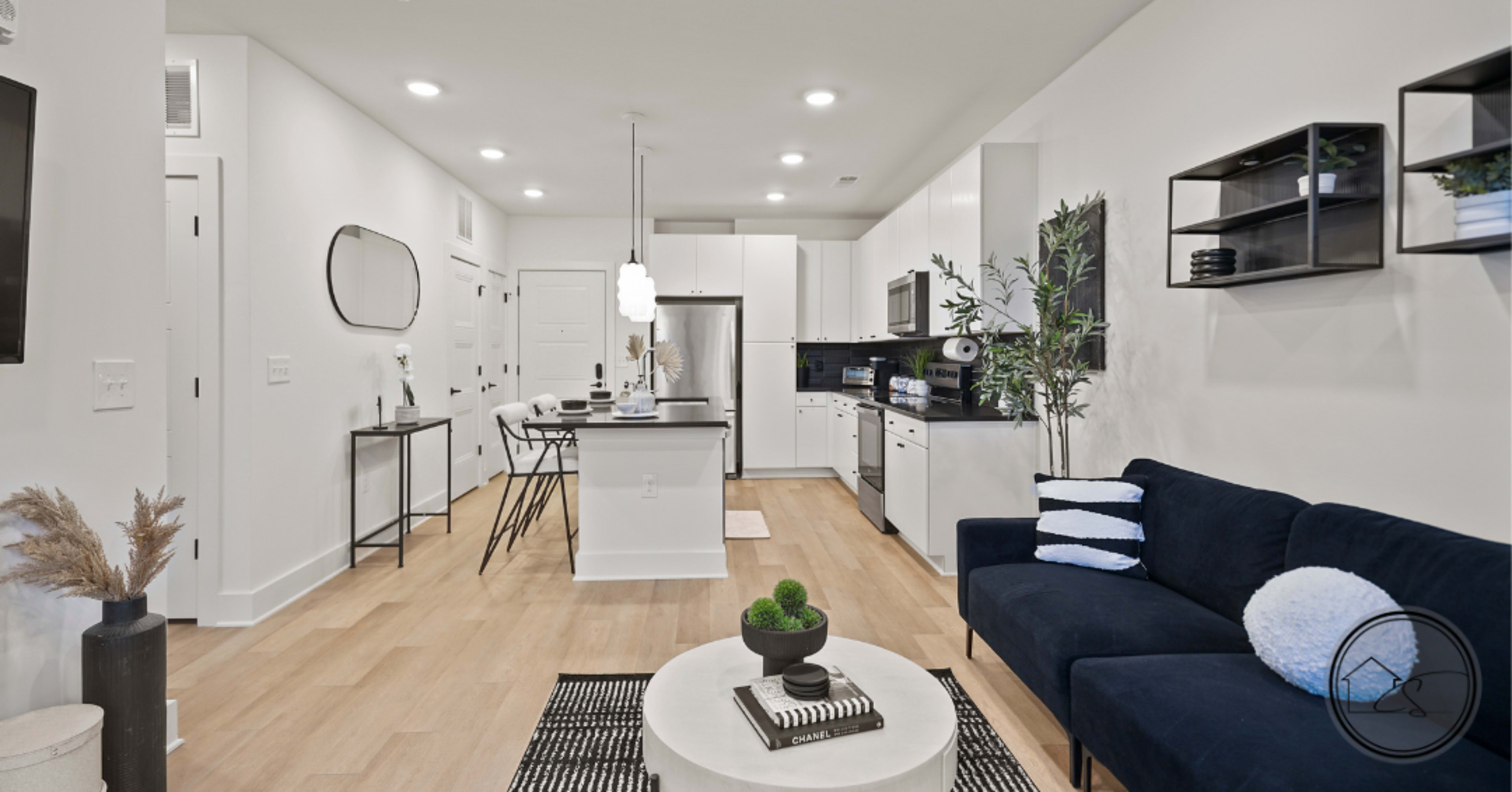 The image shows a modern open-concept kitchen and living area with a dark blue sofa, a round coffee table, and contemporary furnishings.