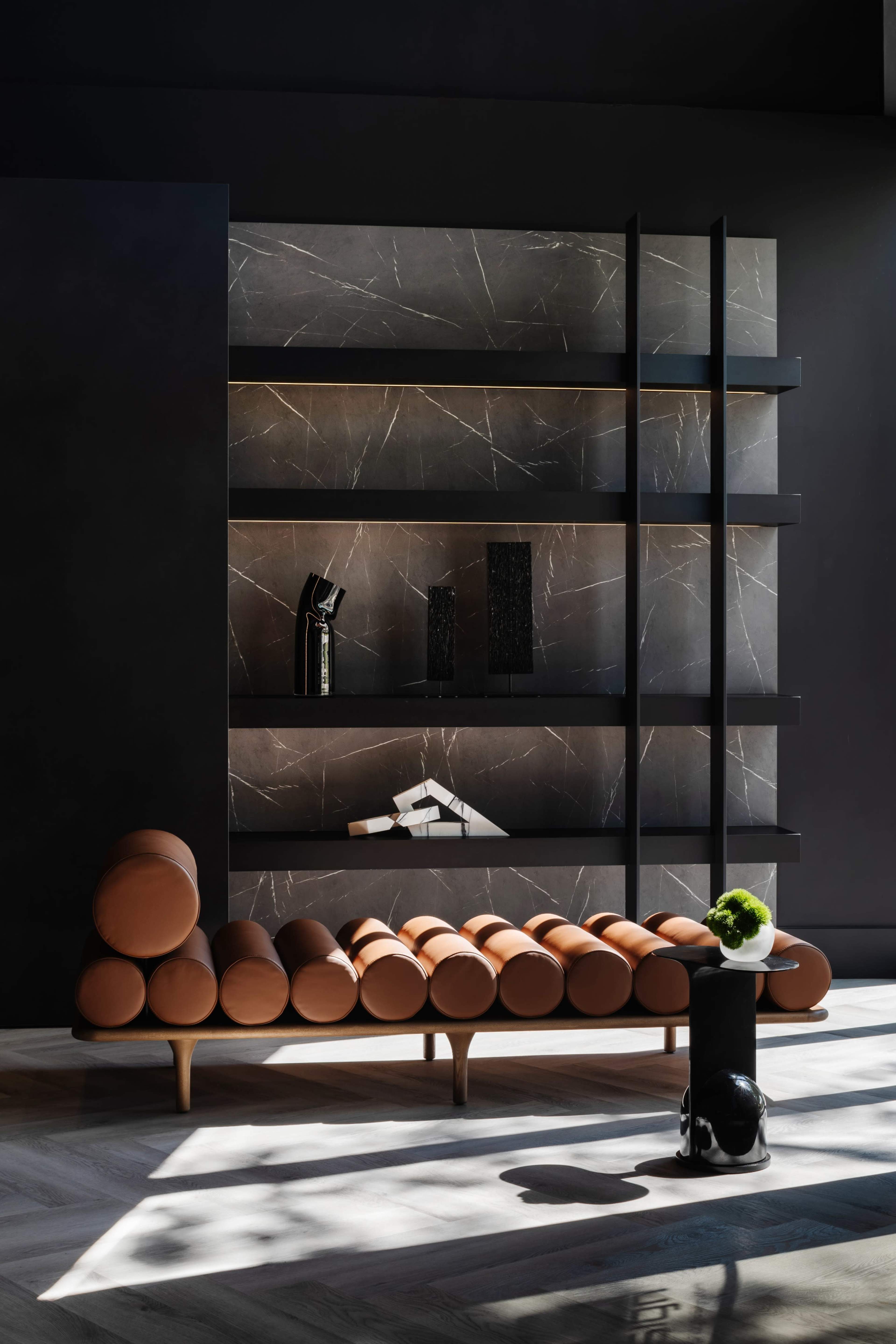 A modern interior featuring a black wall with marble shelves displaying decorative objects, alongside a low, curved couch made of round cushions.