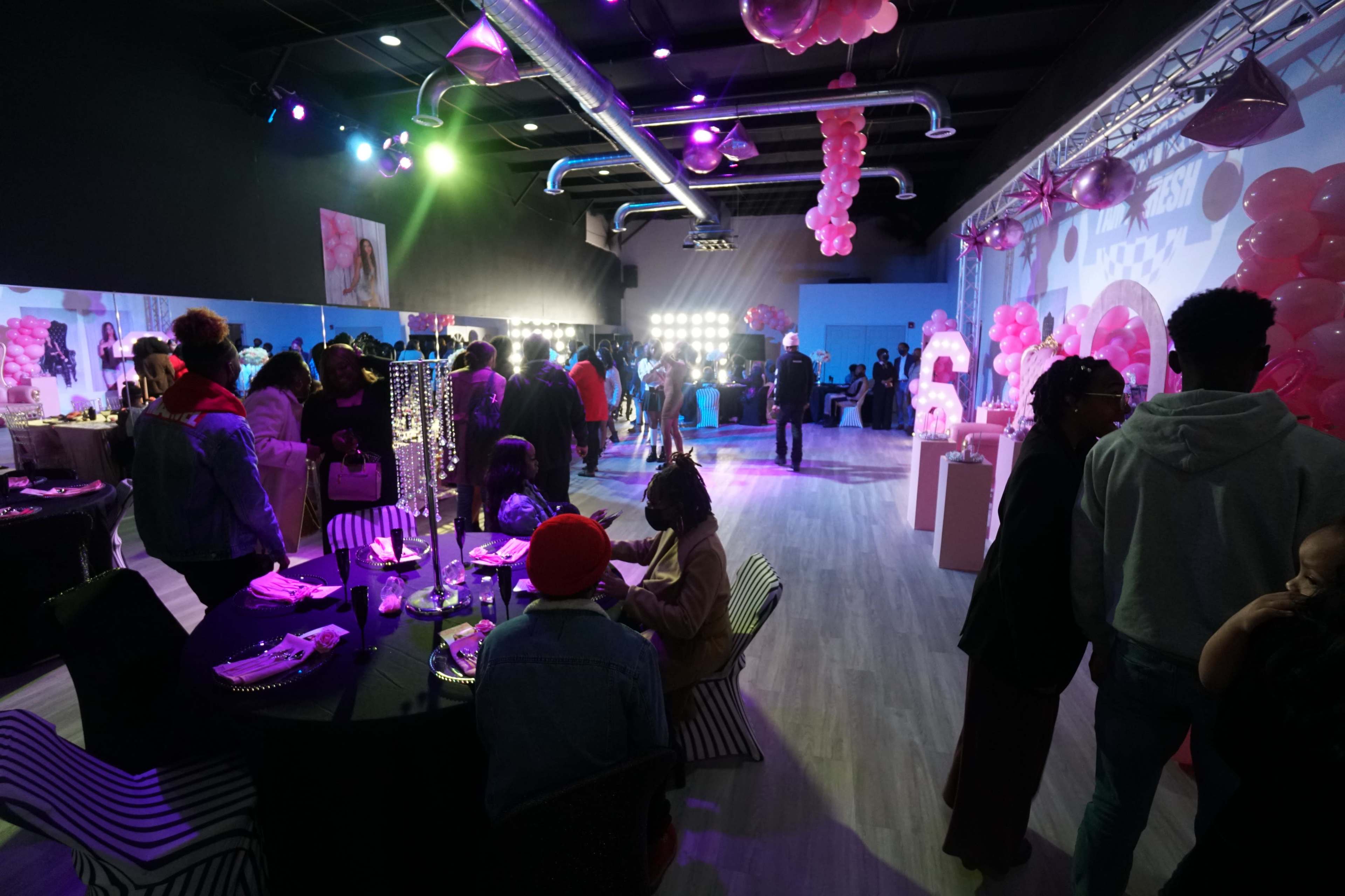 A lively social event is taking place in a decorated indoor space, featuring colorful balloons, lighting effects, and groups of people mingling and enjoying the atmosphere.