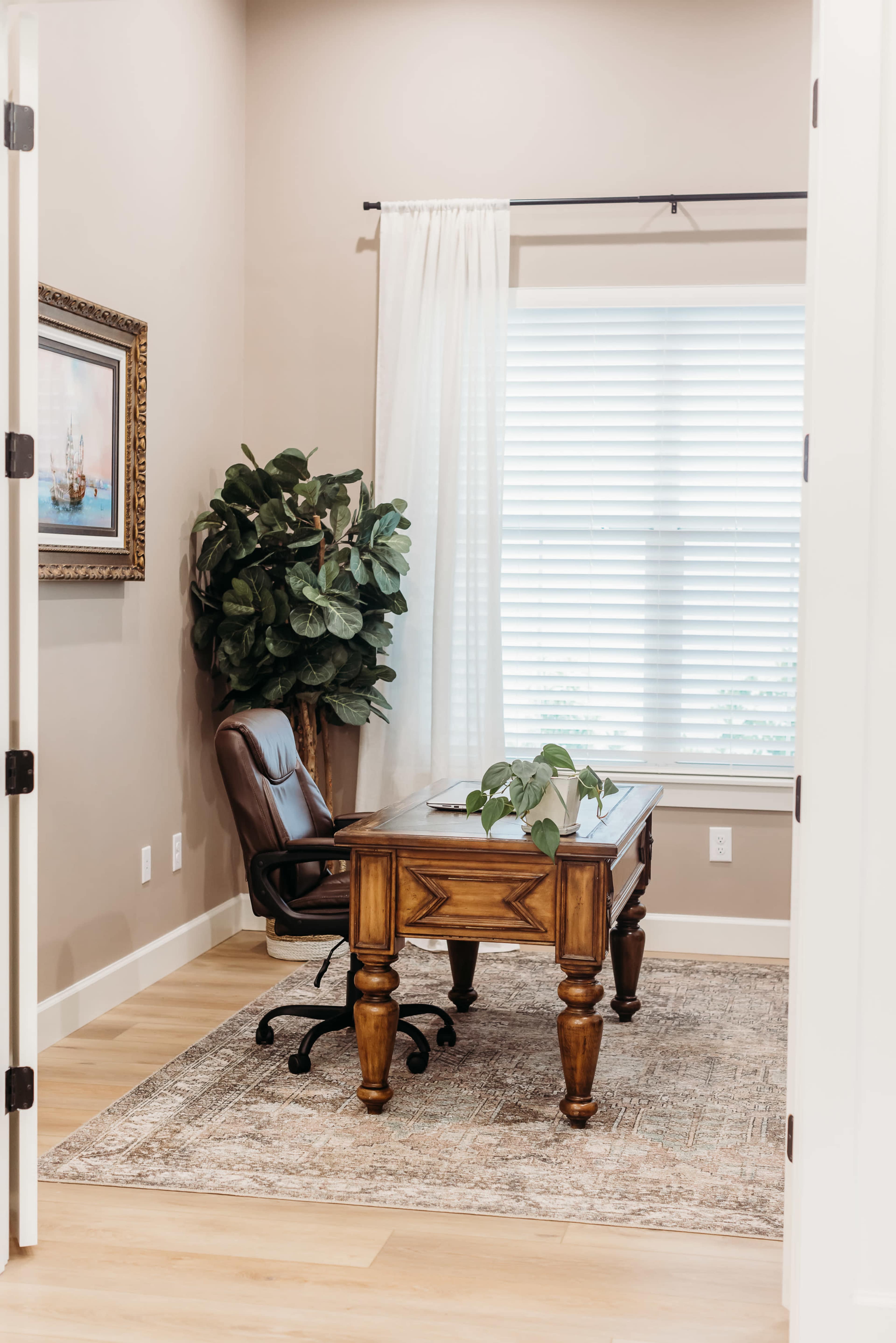 A well-lit office features a wooden desk and a leather chair beside a large window with white blinds and a potted plant.