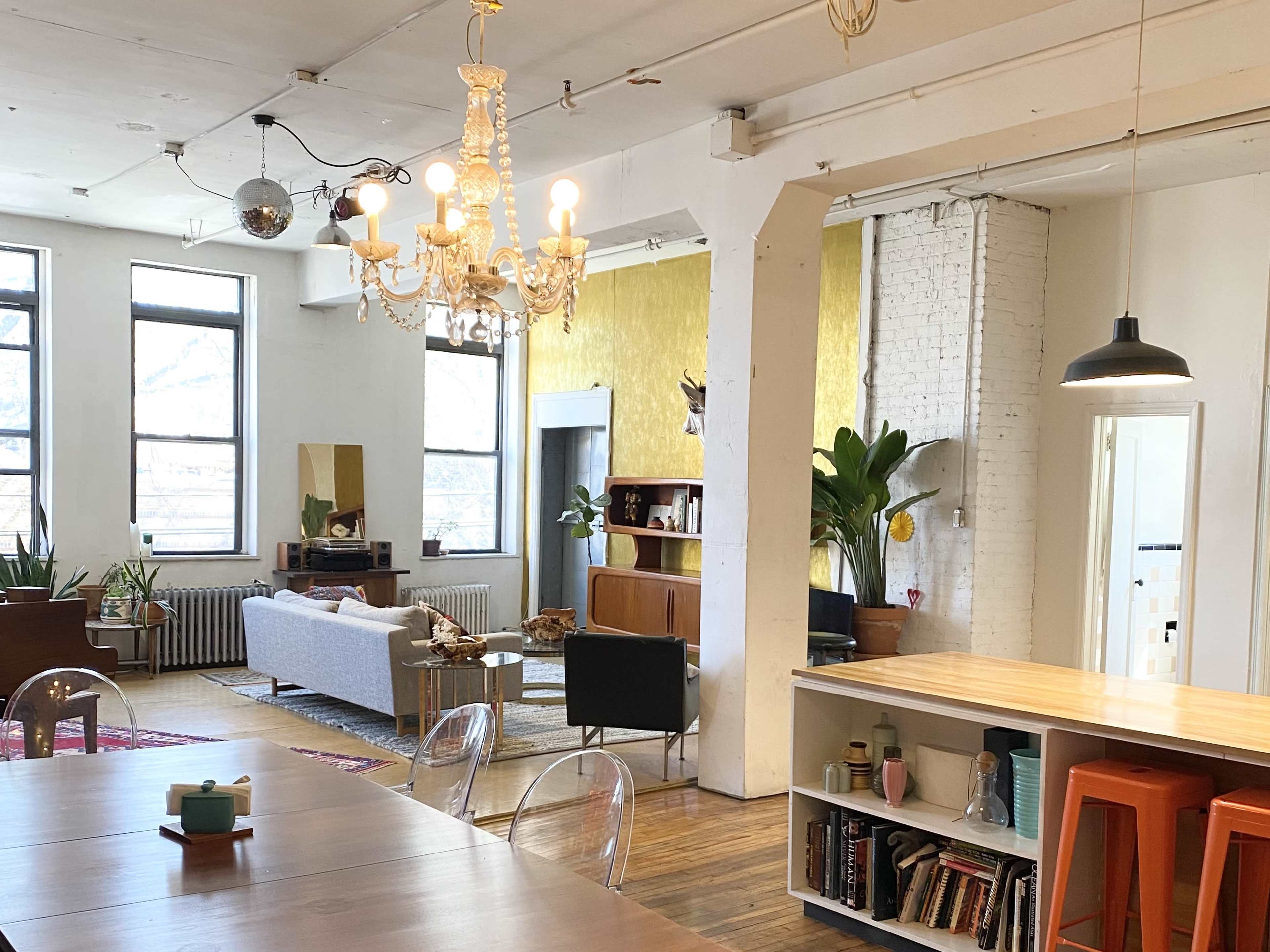 An open, airy loft space with large windows, a dining table, a chandelier, and a cozy living area featuring plants and modern furniture.