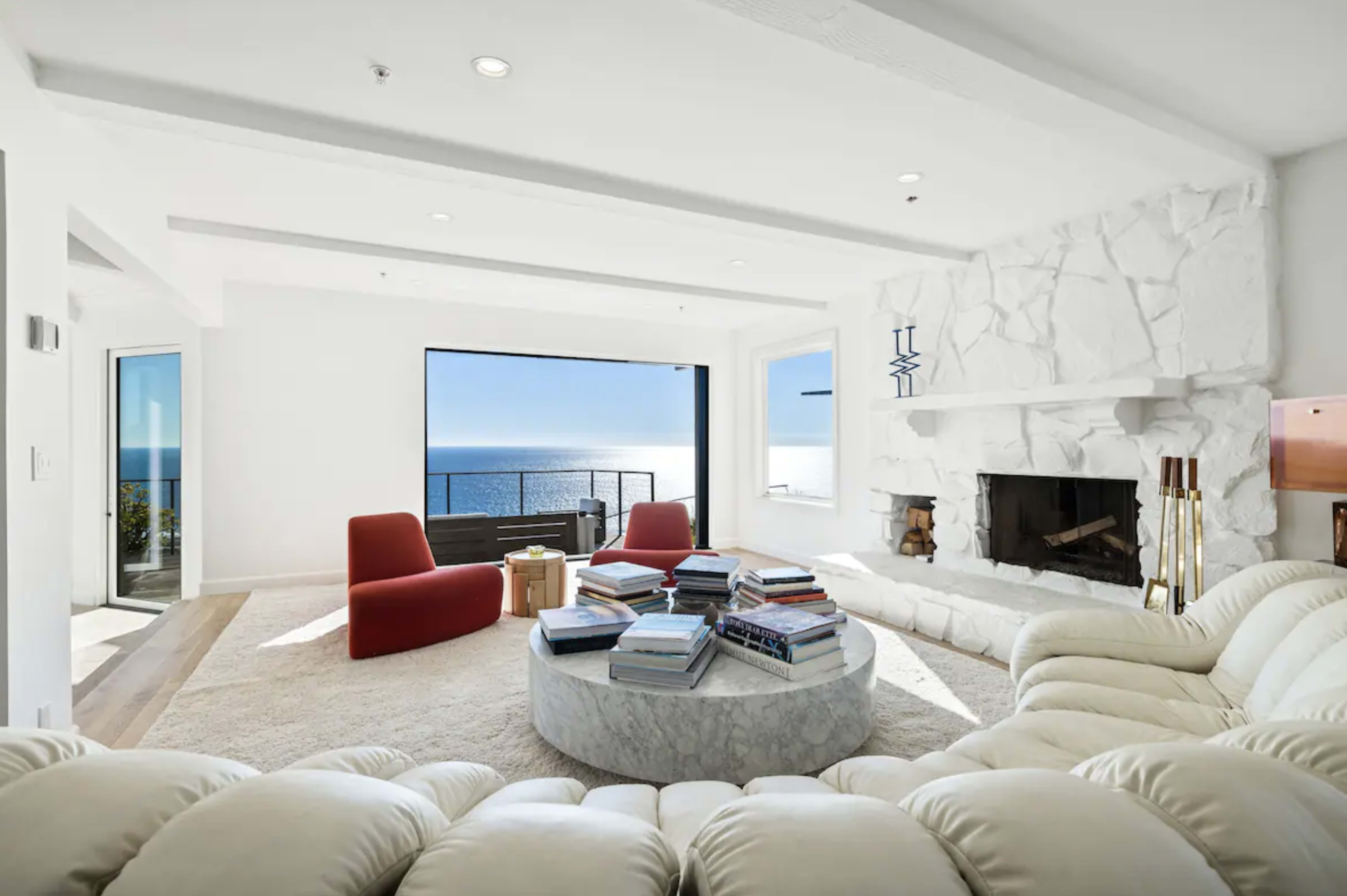 The room features a circular marble coffee table surrounded by a white sectional sofa and red chairs, with large windows showcasing an ocean view.