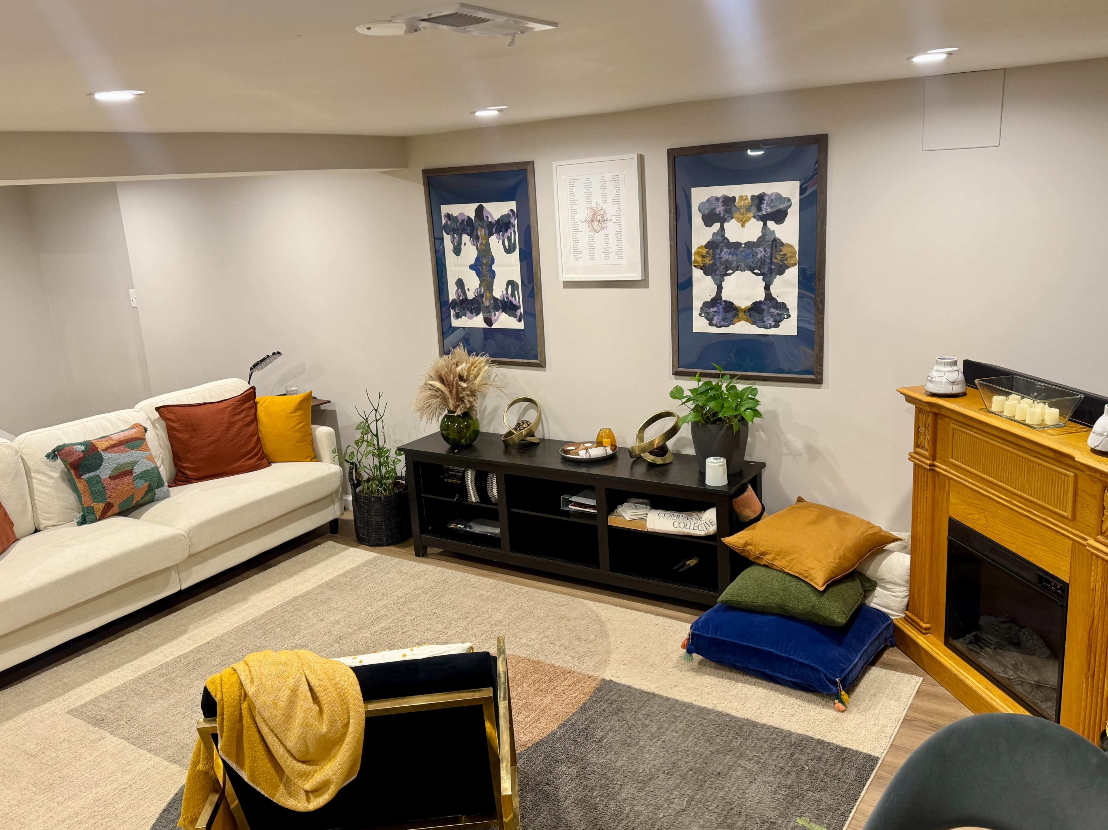 A cozy living room features a sectional sofa with colorful pillows, two framed artworks on the wall, and a wooden fireplace, complemented by plants and various decorative items.