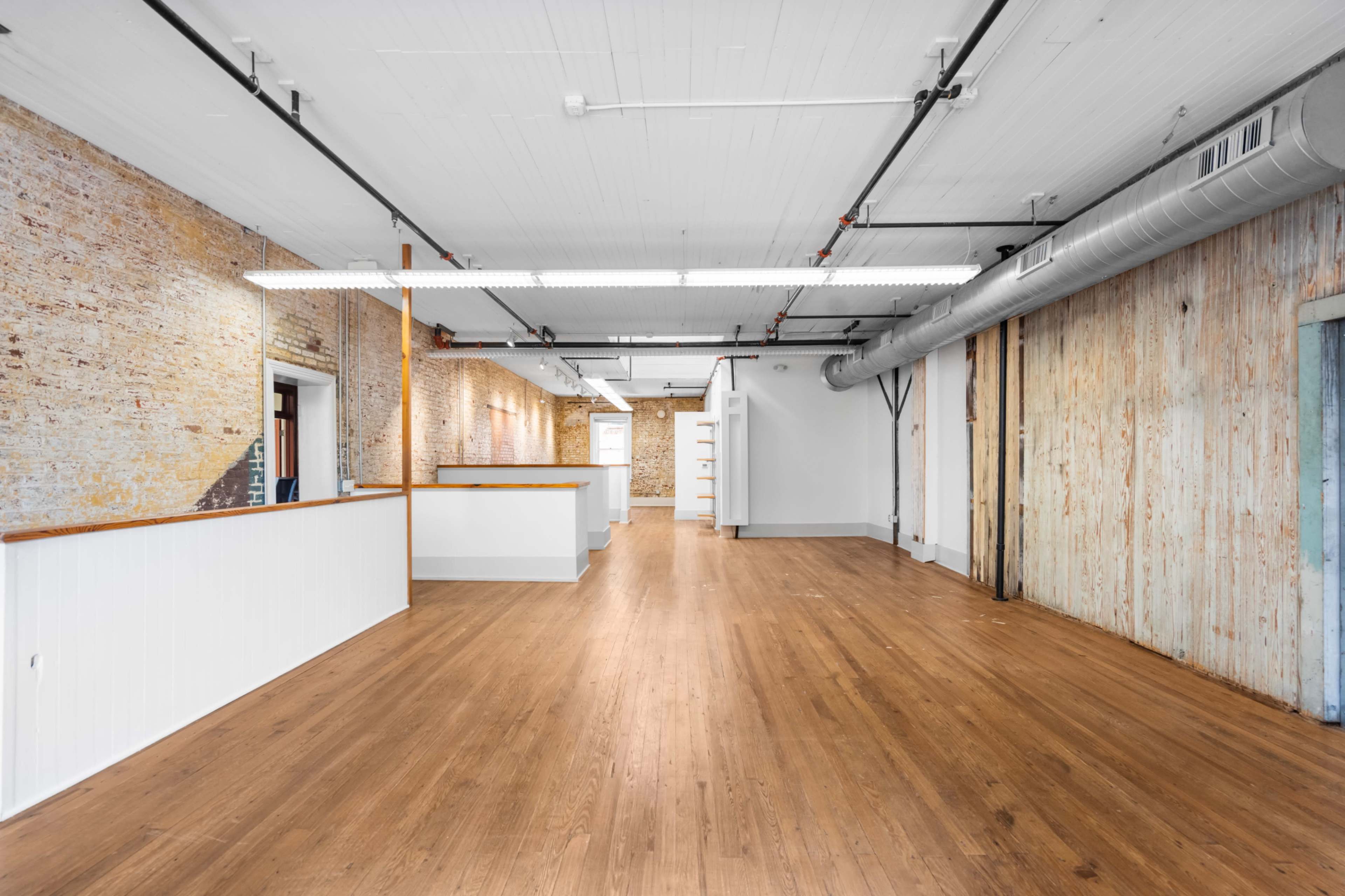 The image shows an empty office space with wooden floors and exposed brick walls, featuring minimal partitions and overhead lighting.