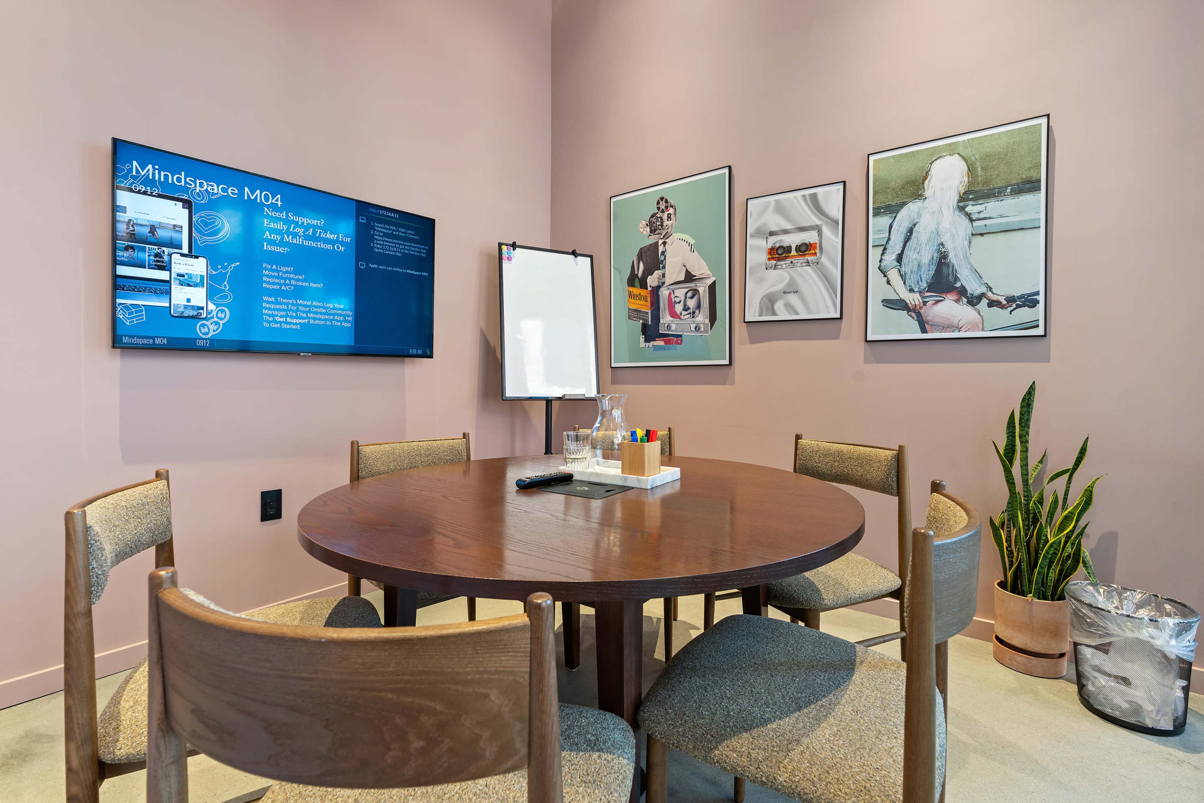 A modern meeting room with a round wooden table, four chairs, a digital screen displaying information, a whiteboard, art on the walls, and a potted plant in the corner.