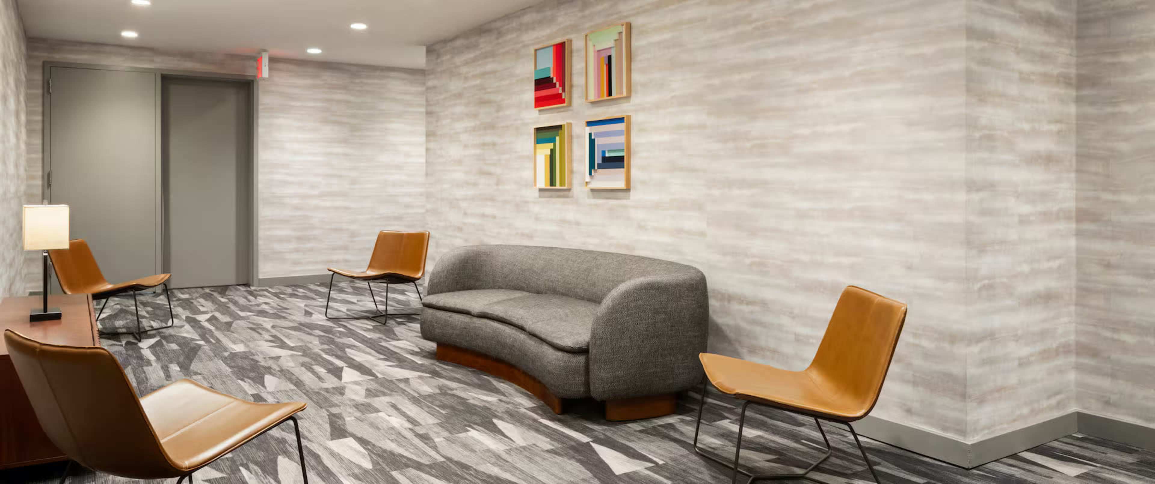 The image shows a modern waiting area with a gray upholstered sofa and four tan chairs, featuring abstract art on the wall.