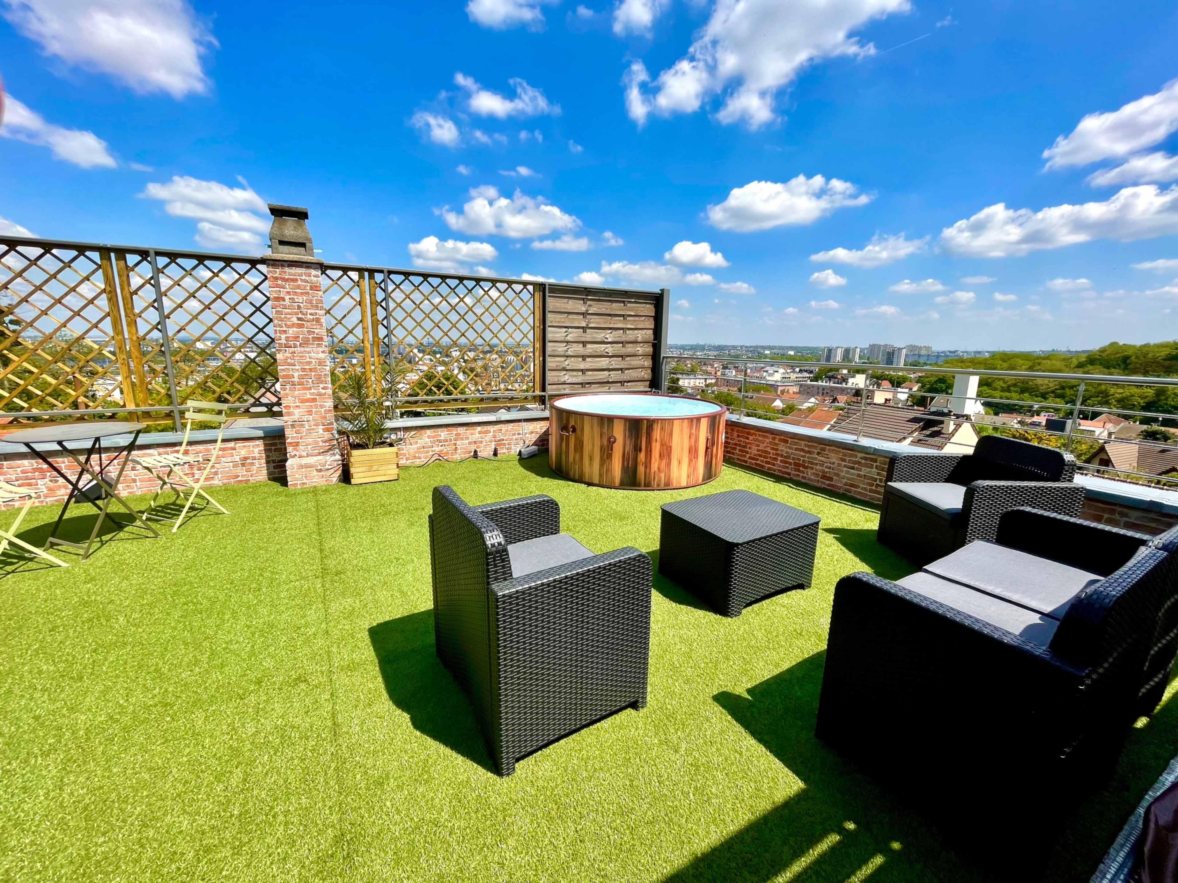 A rooftop terrace features a wooden hot tub, black wicker chairs, and a small table on a grassy surface under a blue sky with clouds.