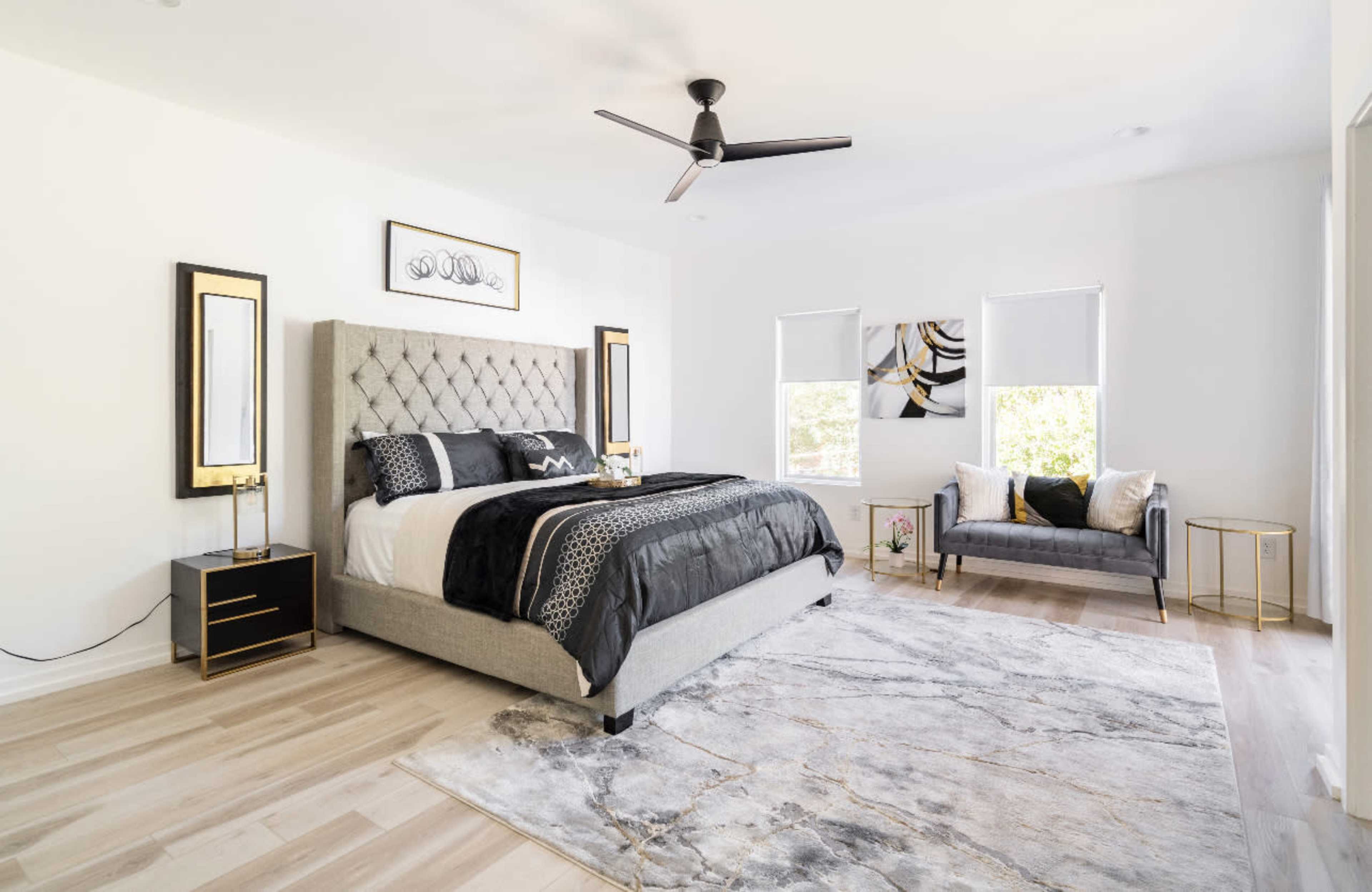 A modern bedroom with a gray upholstered bed, a large area rug, and two windows featuring light-colored shades.