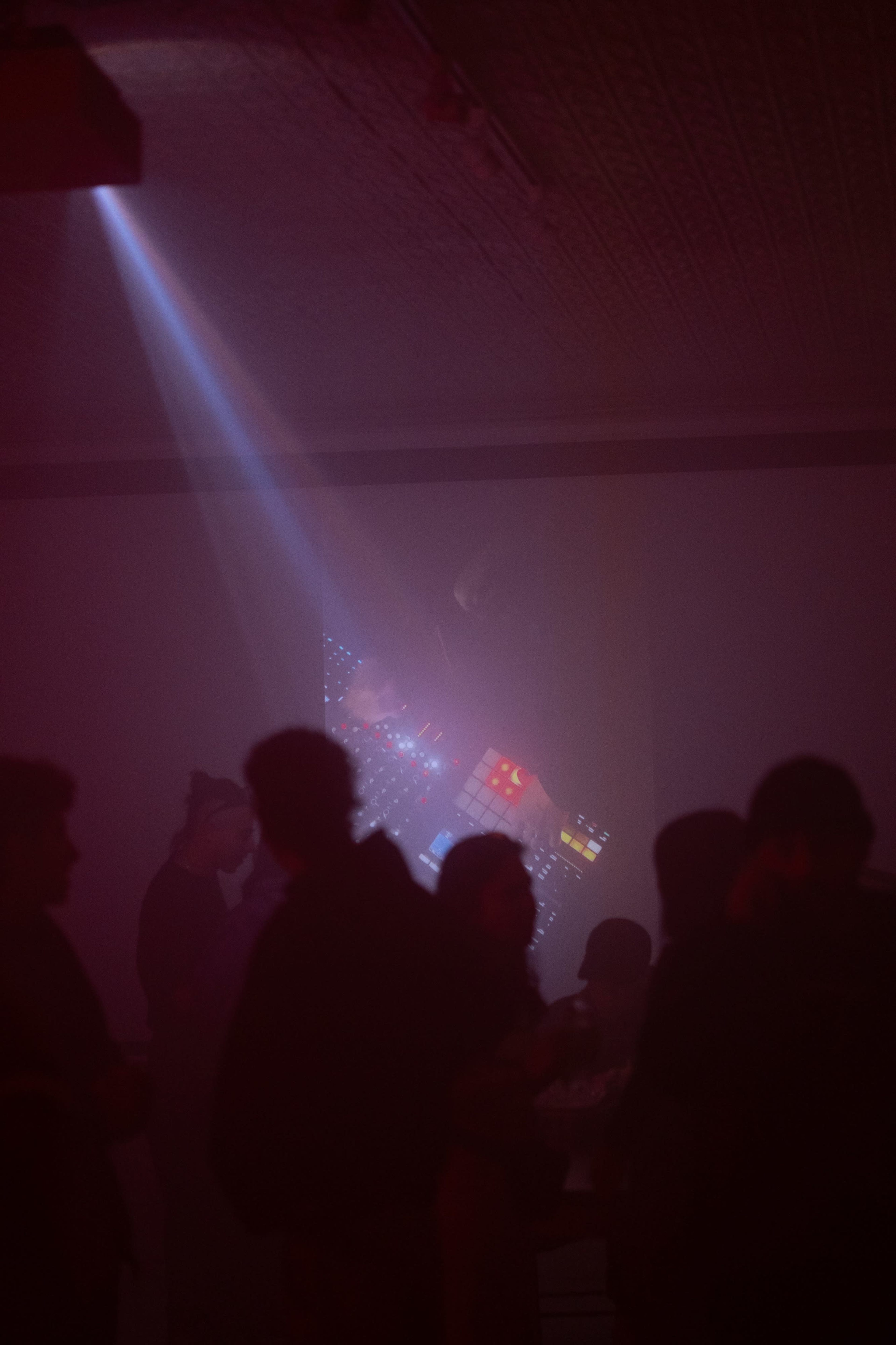 A group of people gather in a dimly lit room with a projection of colorful lights and patterns on the wall.
