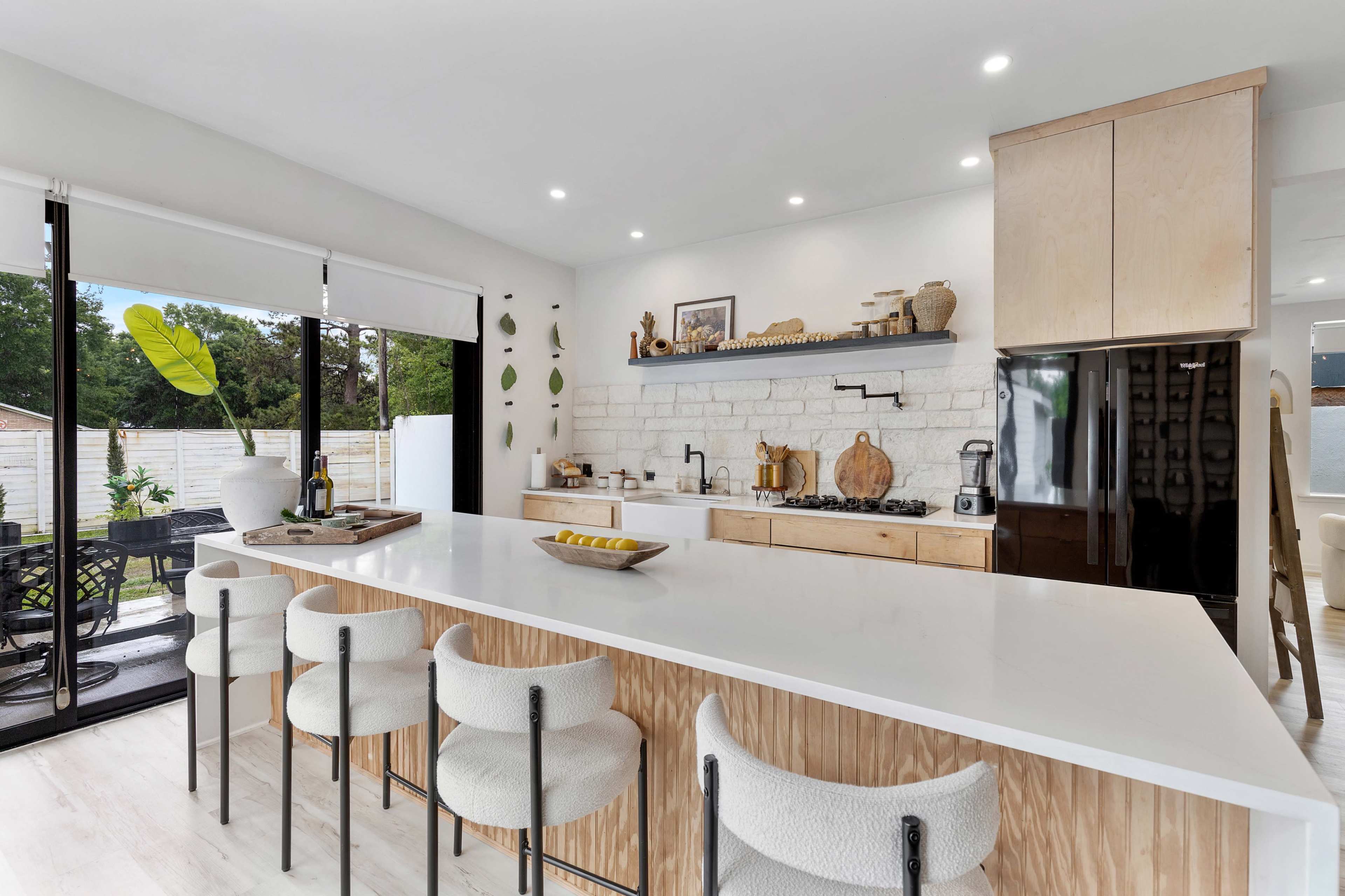 The image shows a modern kitchen with a large island, white cabinetry, and a view of an outdoor seating area through glass doors.