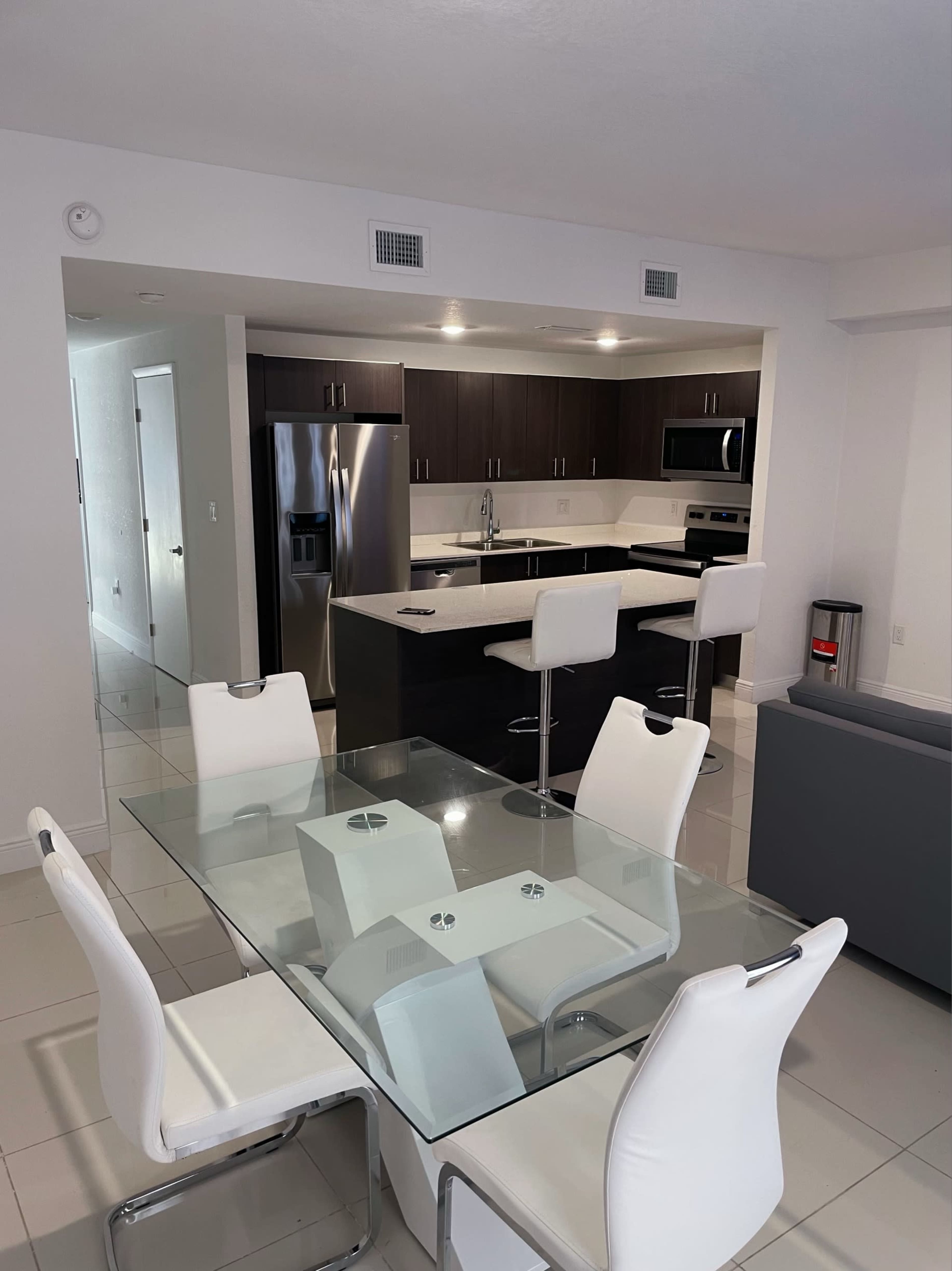 A modern kitchen with dark cabinetry and stainless steel appliances is visible from a dining area featuring a glass table and white chairs.