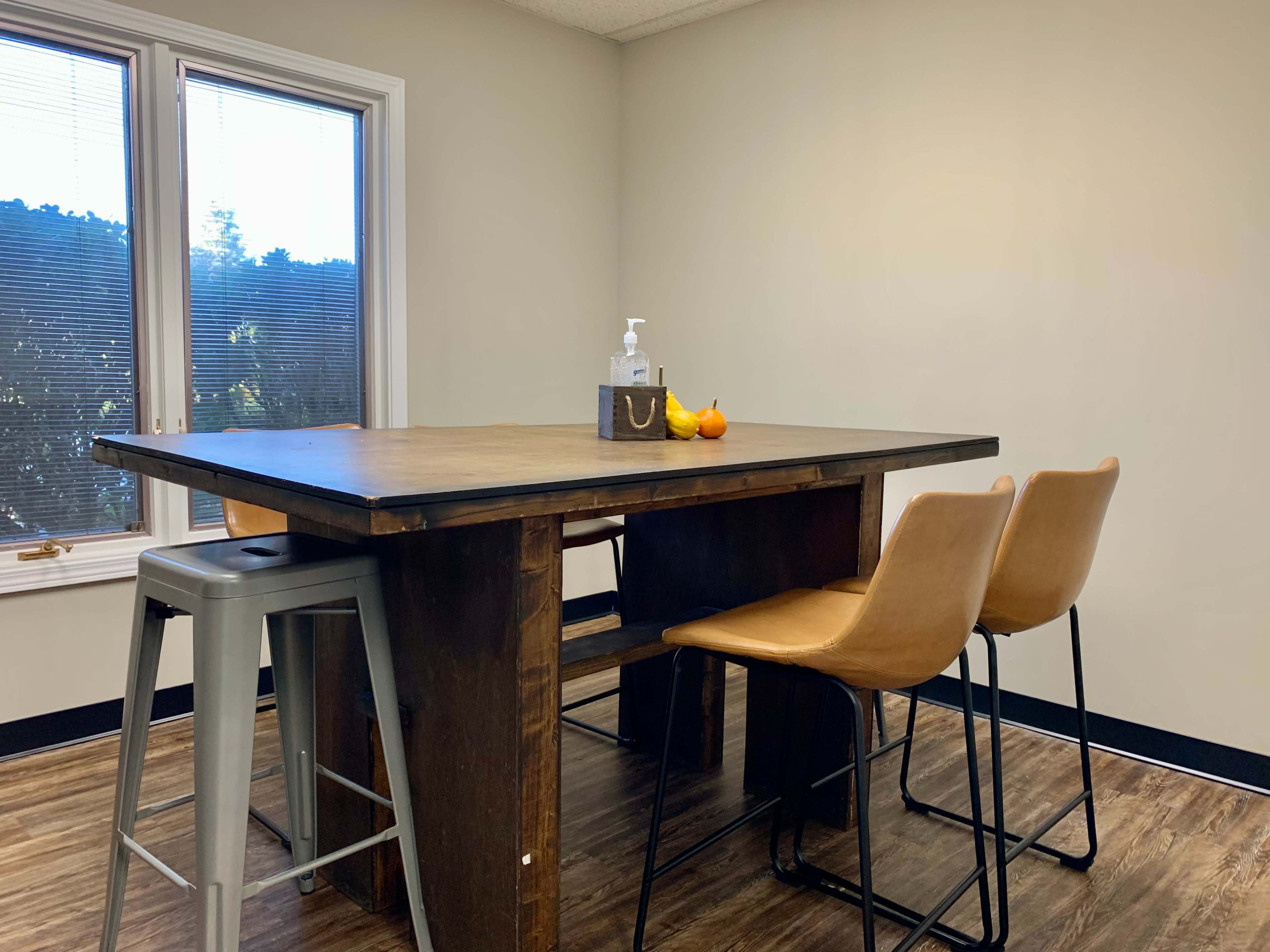 A wooden table with three leather stools is set near a window, featuring a hand sanitizer dispenser and a few pieces of fruit on top.