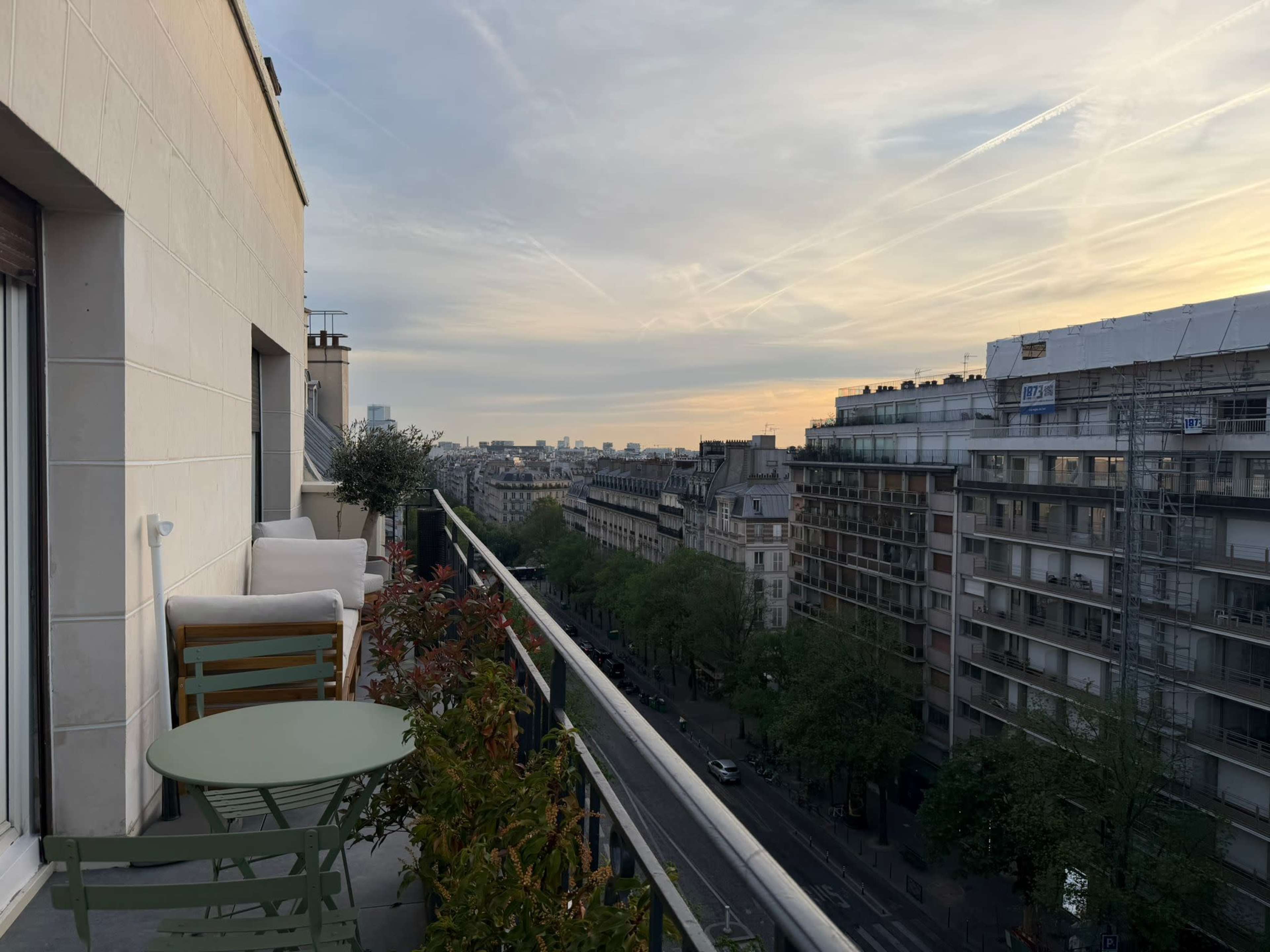 The image shows a balcony with a small green table and chairs overlooking a cityscape at sunset.