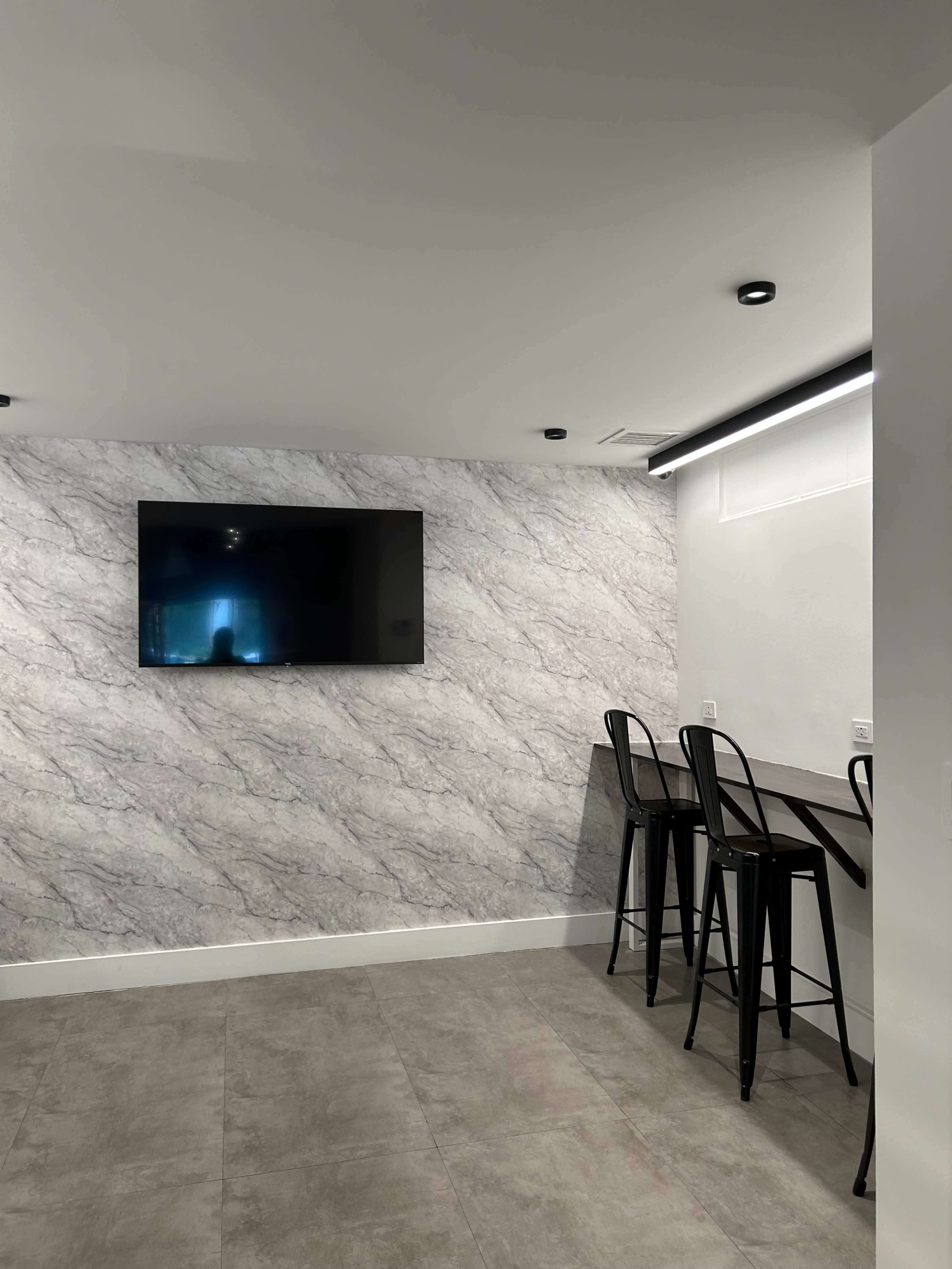 The image shows a modern interior space featuring a flat-screen TV mounted on a marbled wall and a bar-style seating area with black stools.