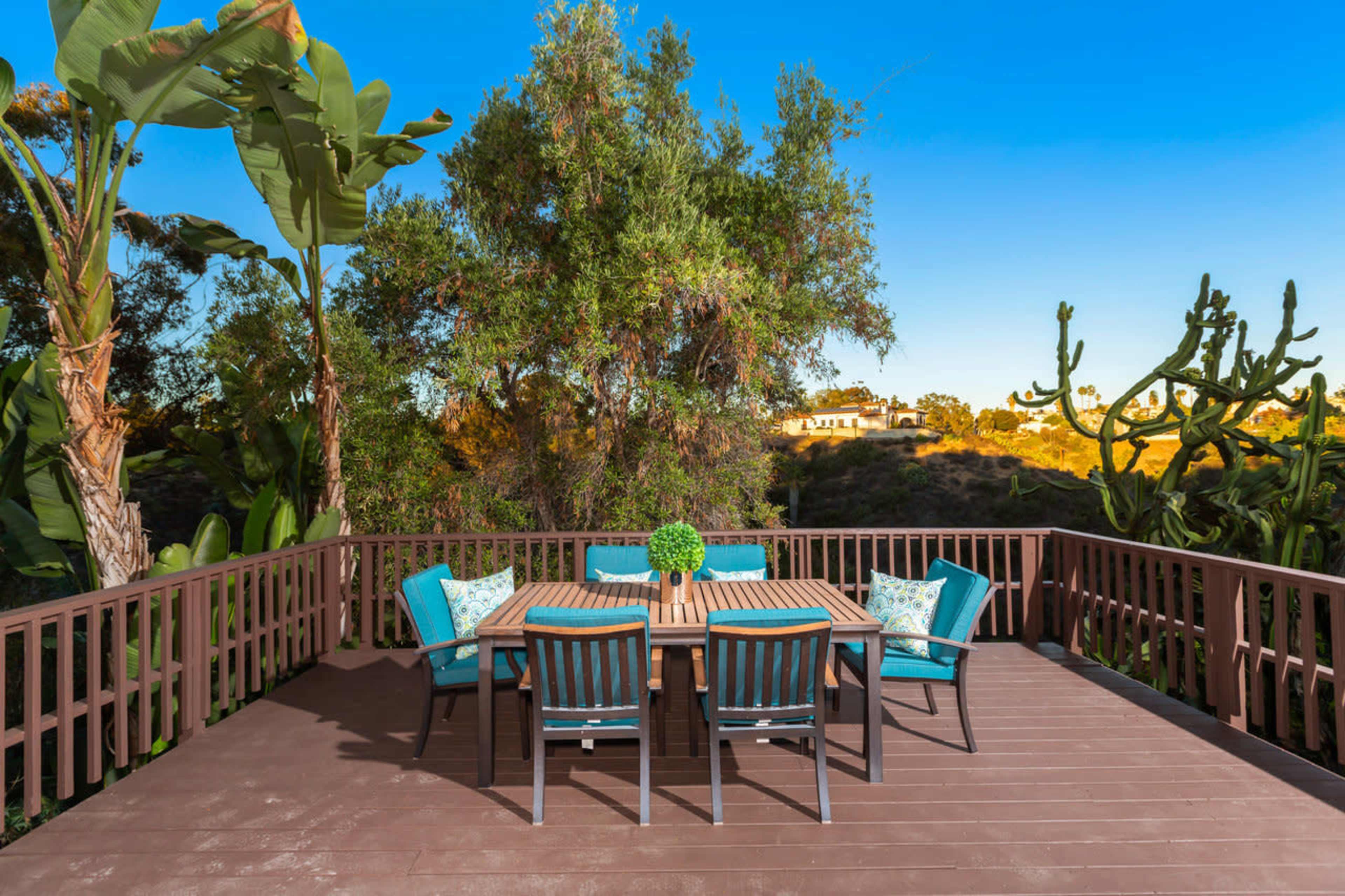 A wooden deck with a dining table and six chairs is surrounded by greenery and cacti in a residential area.
