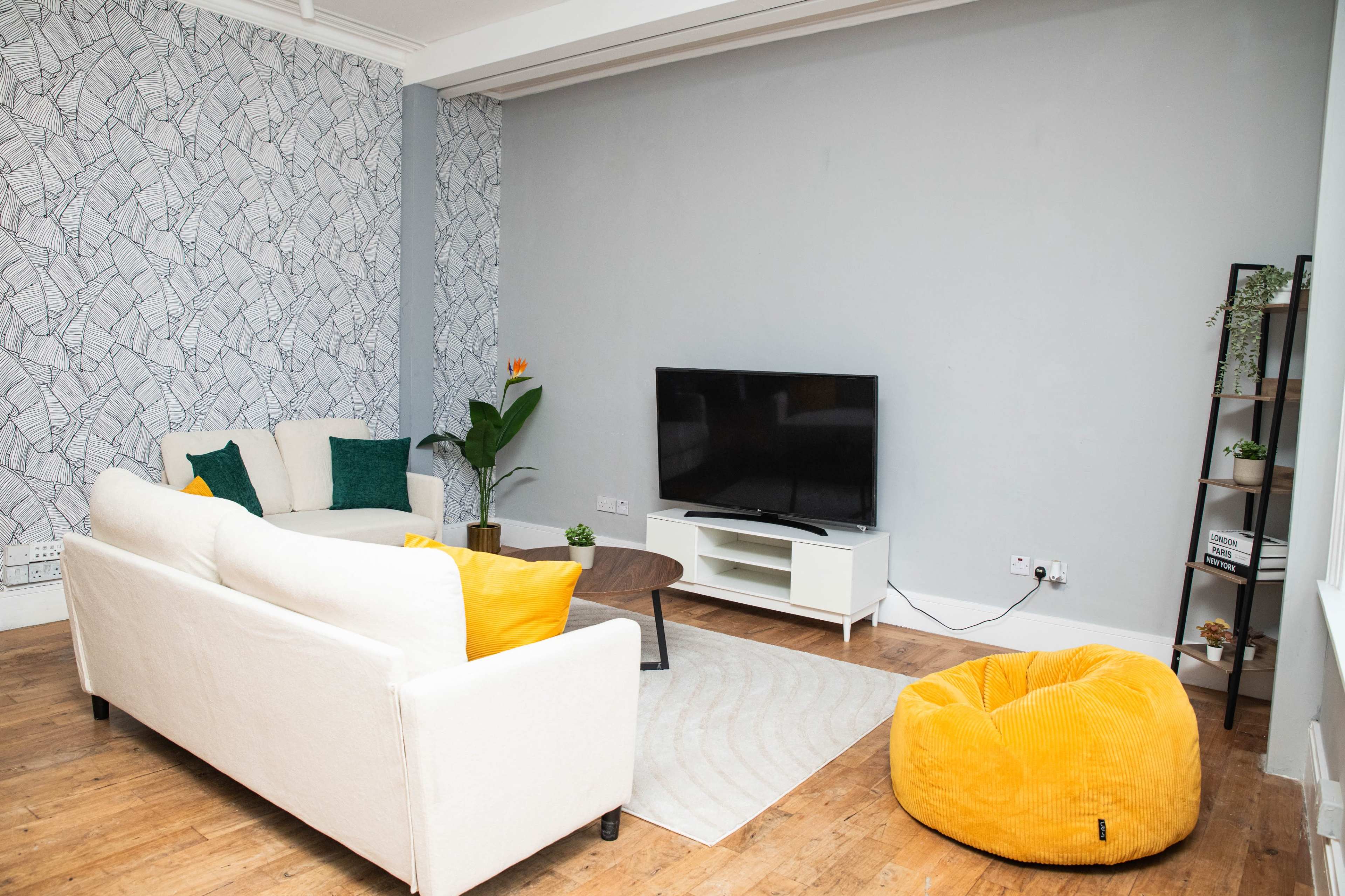 The image shows a modern living room with a white couch, a coffee table, a television on a stand, a yellow bean bag, and a patterned wallpaper.