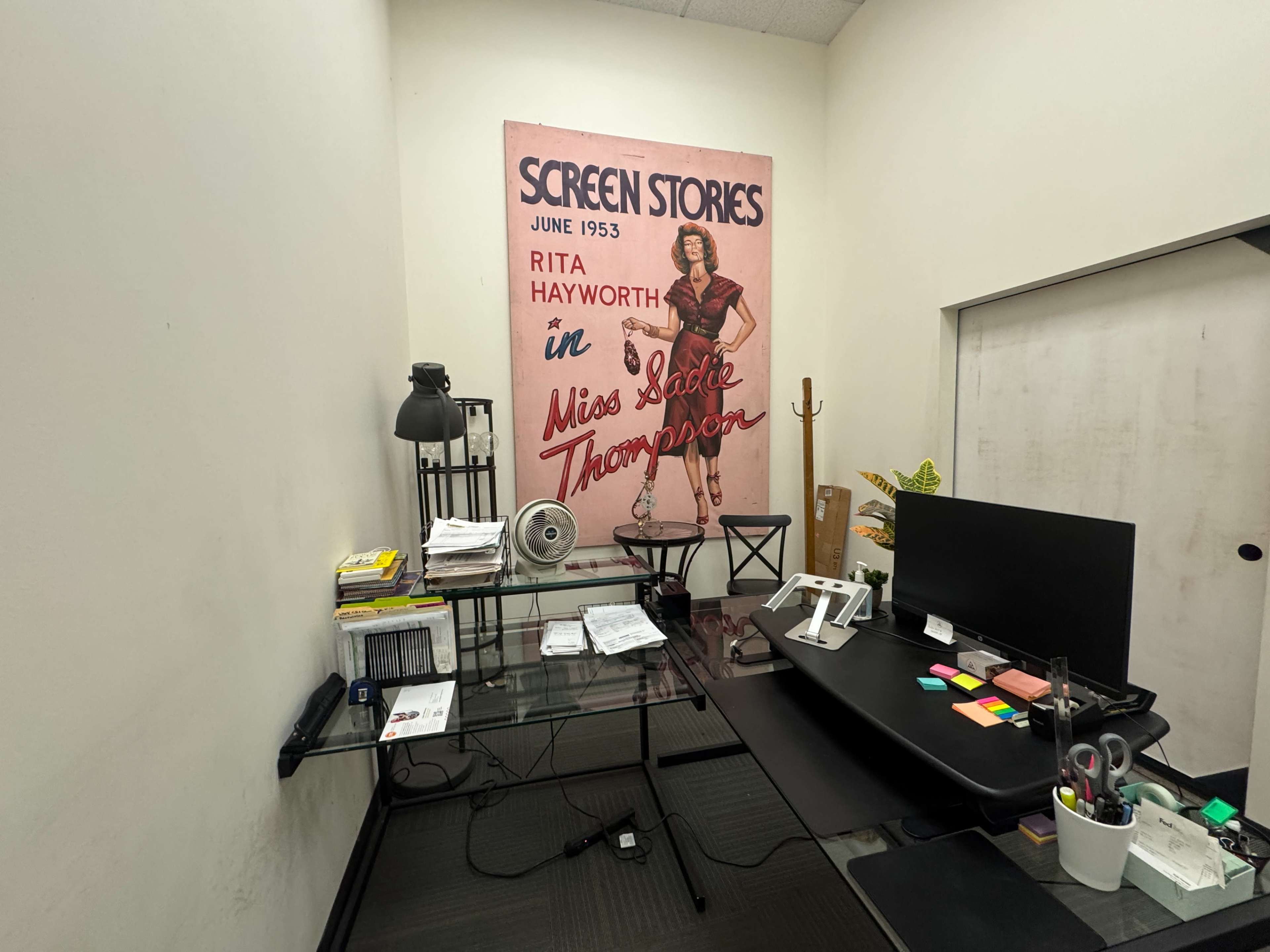 The image shows a small office space featuring a glass desk, a computer monitor, and a large poster of Rita Hayworth from June 1953, titled "Screen Stories."