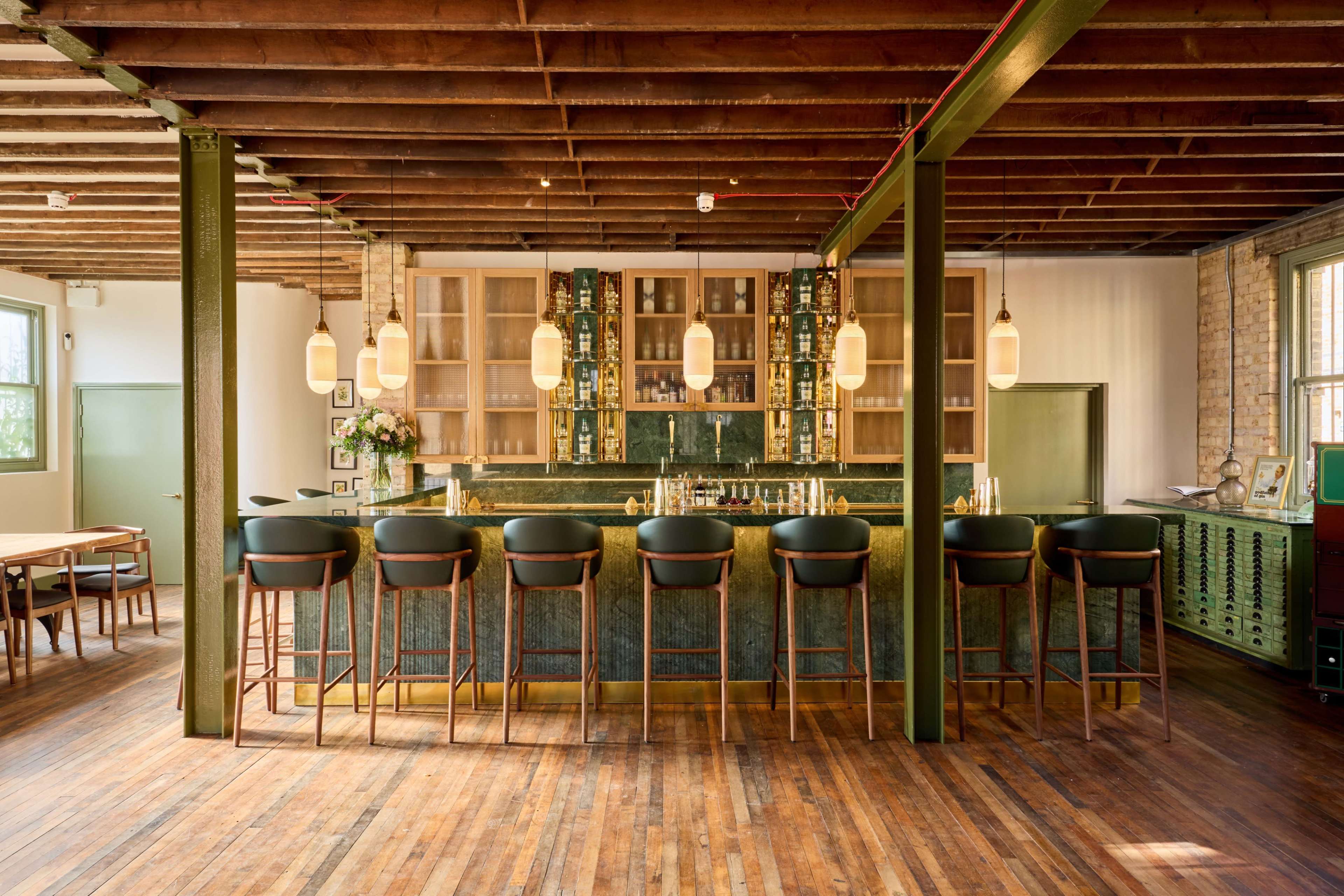 A polished wooden bar with high stools is positioned in a spacious room featuring exposed beams and a display of various bottles behind the counter.