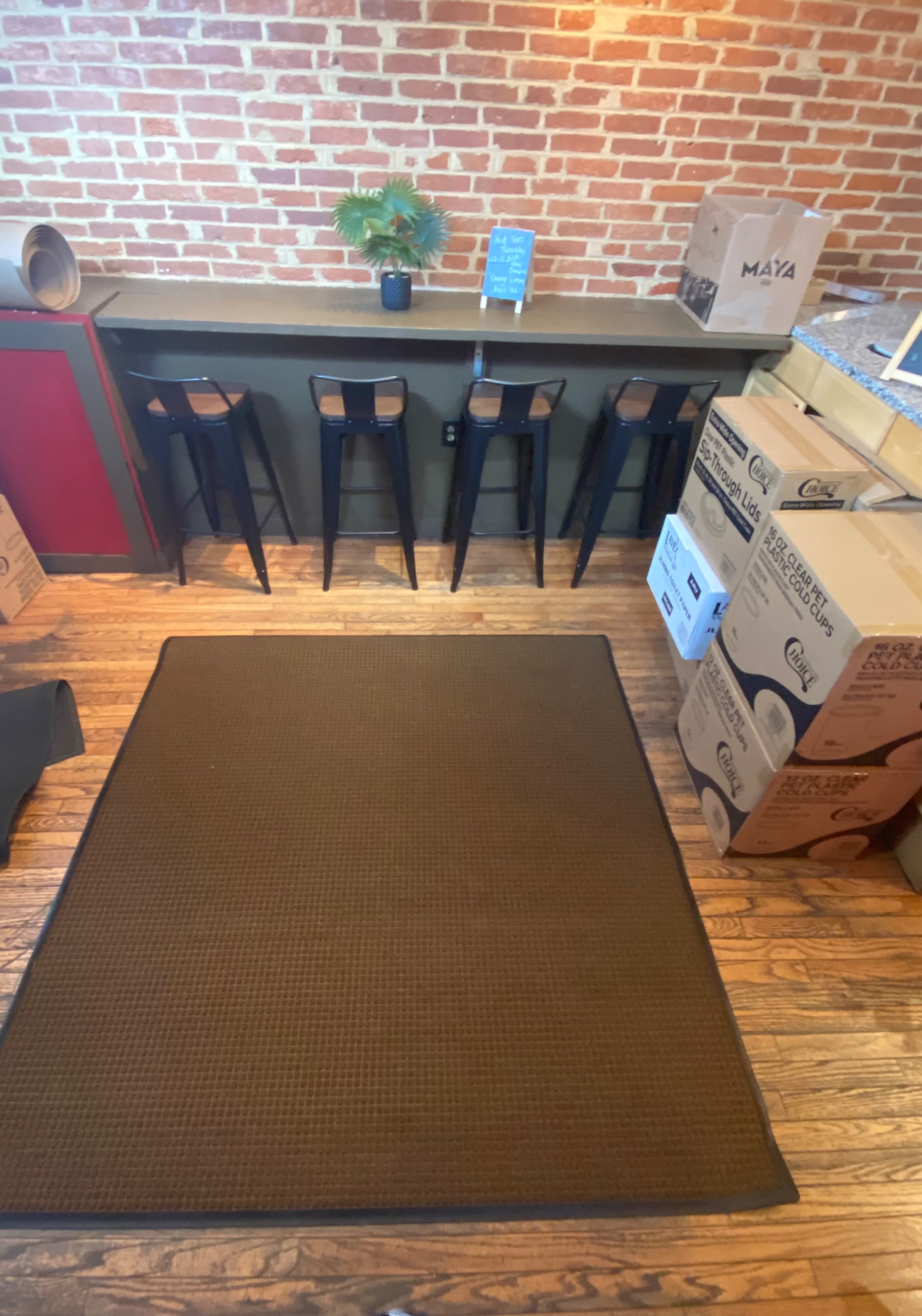 The image shows a cozy space with a brick wall, a bar-like counter with four black stools, and a carpeted area in front, surrounded by unpacked boxes.