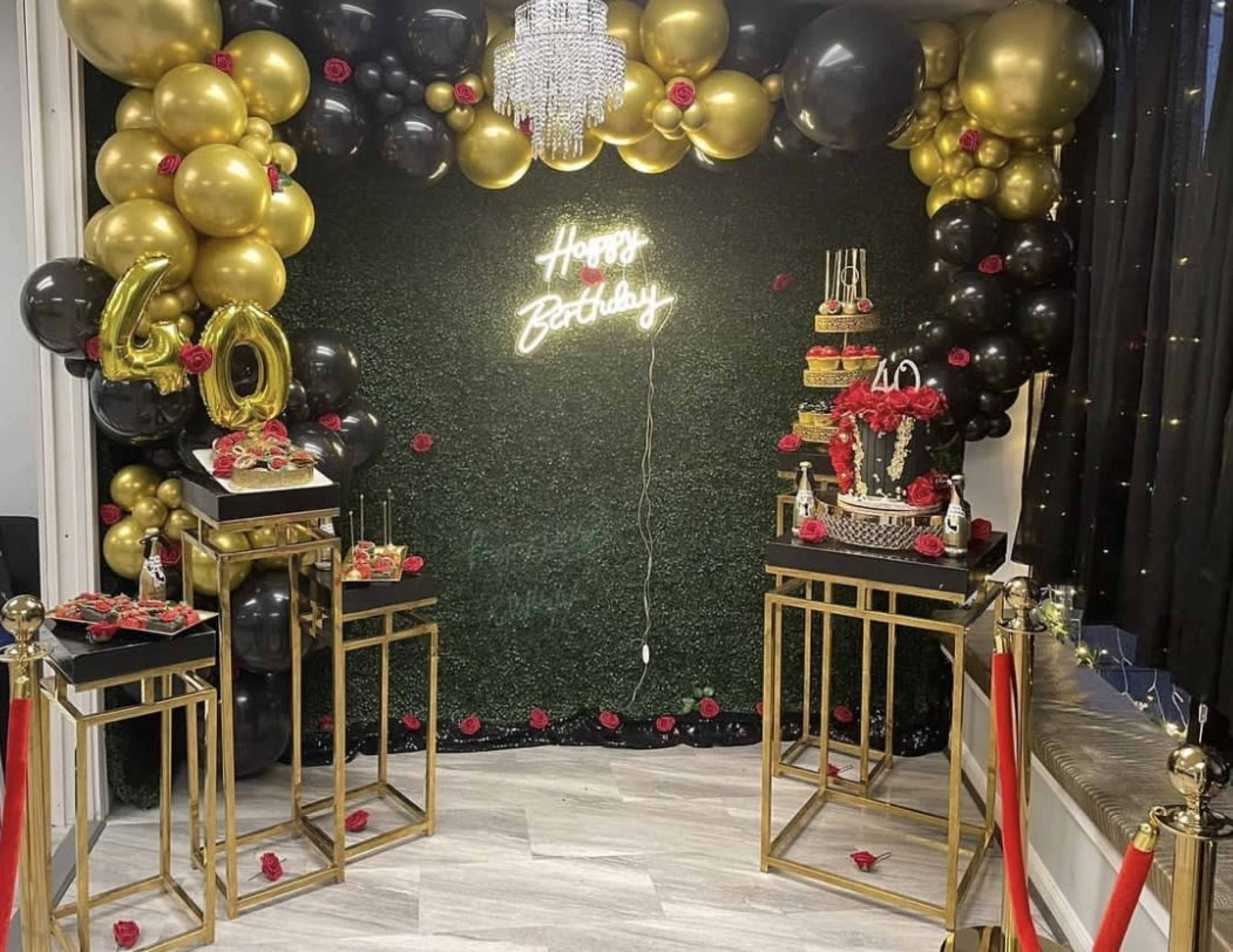 A decorated birthday party setup featuring a neon sign that says "Happy Birthday," a large cake on a stand, and a backdrop of black, gold, and red balloons.