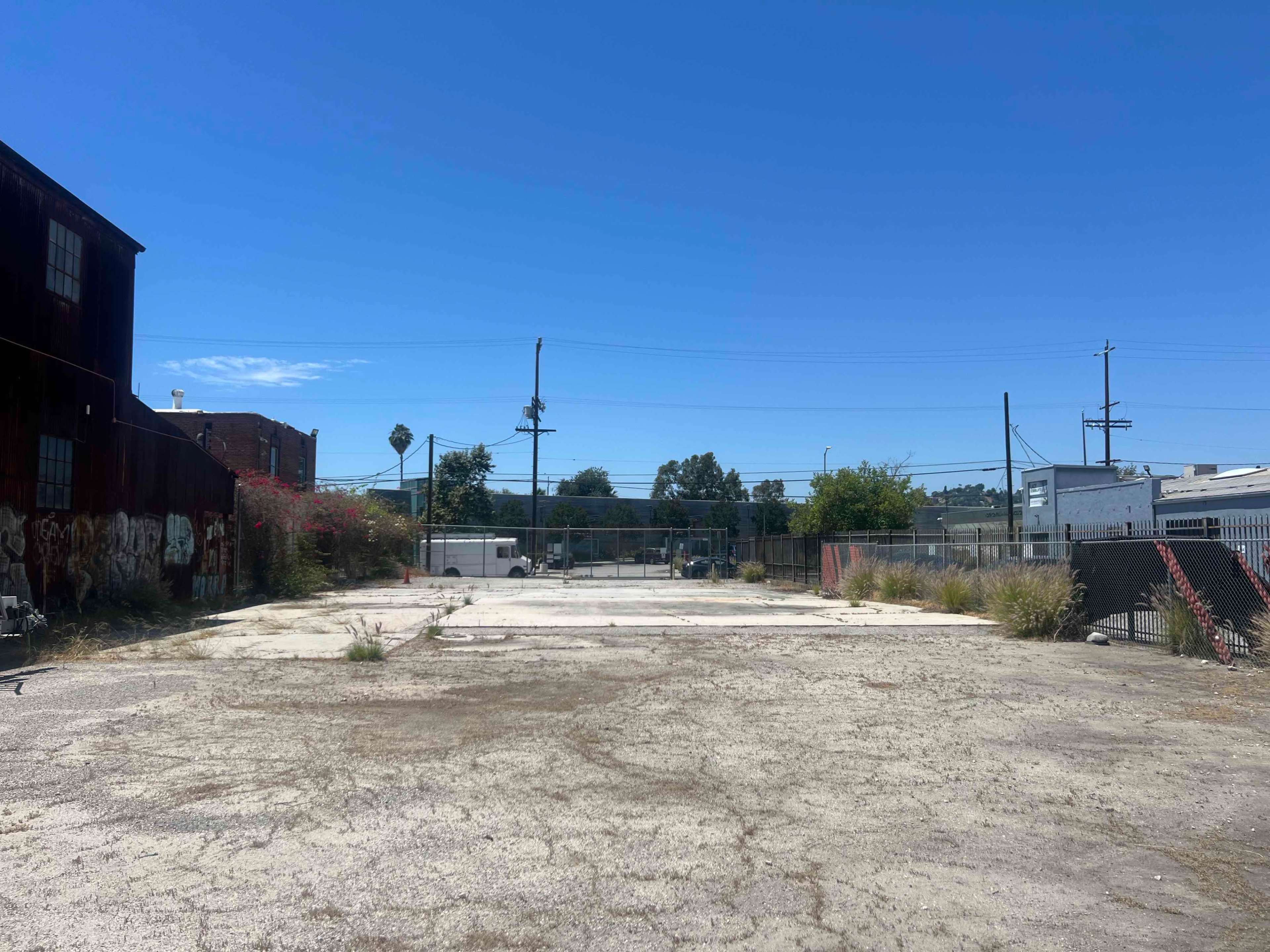 An empty, paved lot surrounded by overgrown weeds, with buildings and power lines in the background under a clear blue sky.