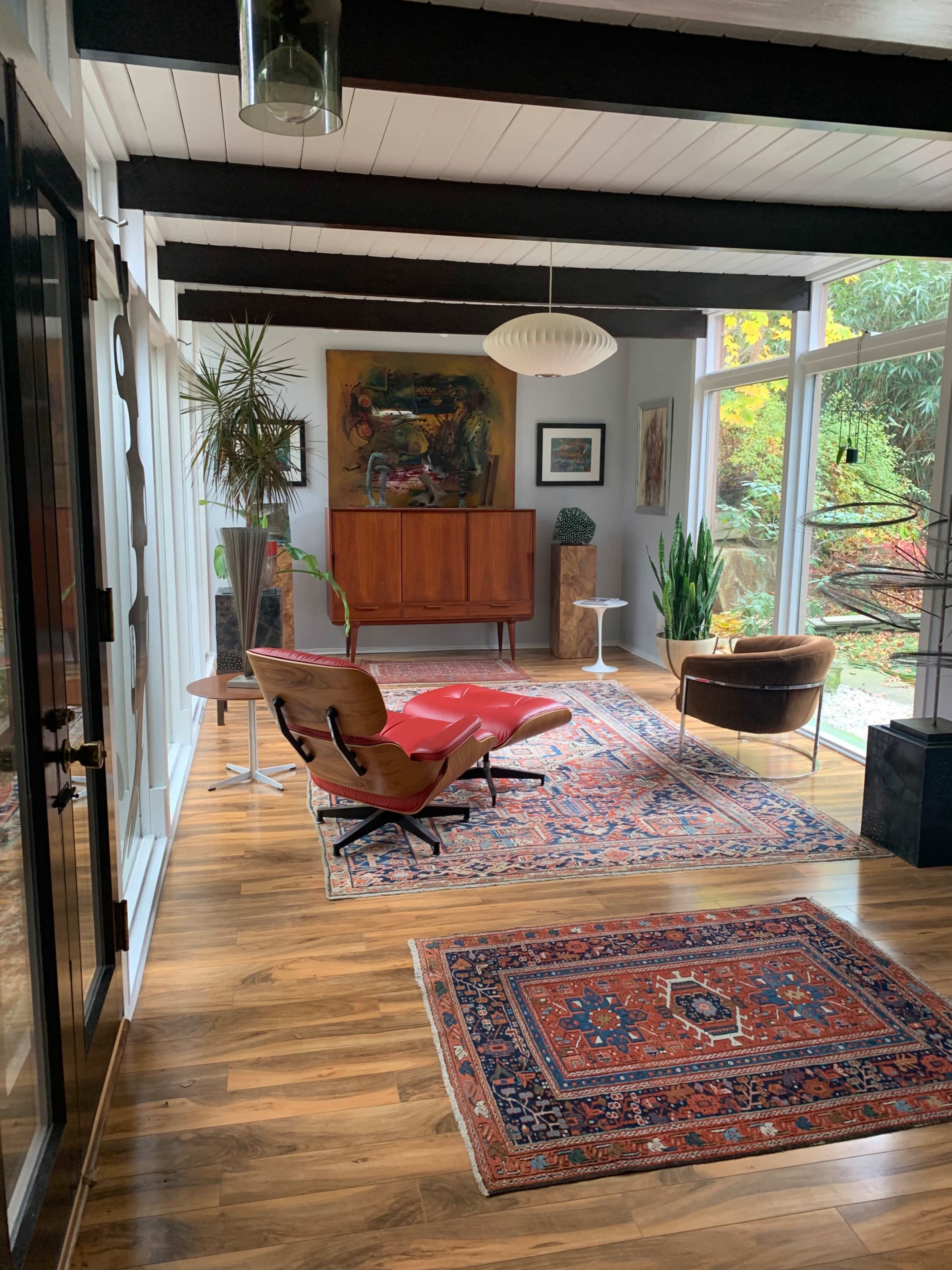 The image shows a well-lit interior space featuring a mid-century modern lounge area with a red chair, a wooden sideboard, and large windows overlooking greenery.