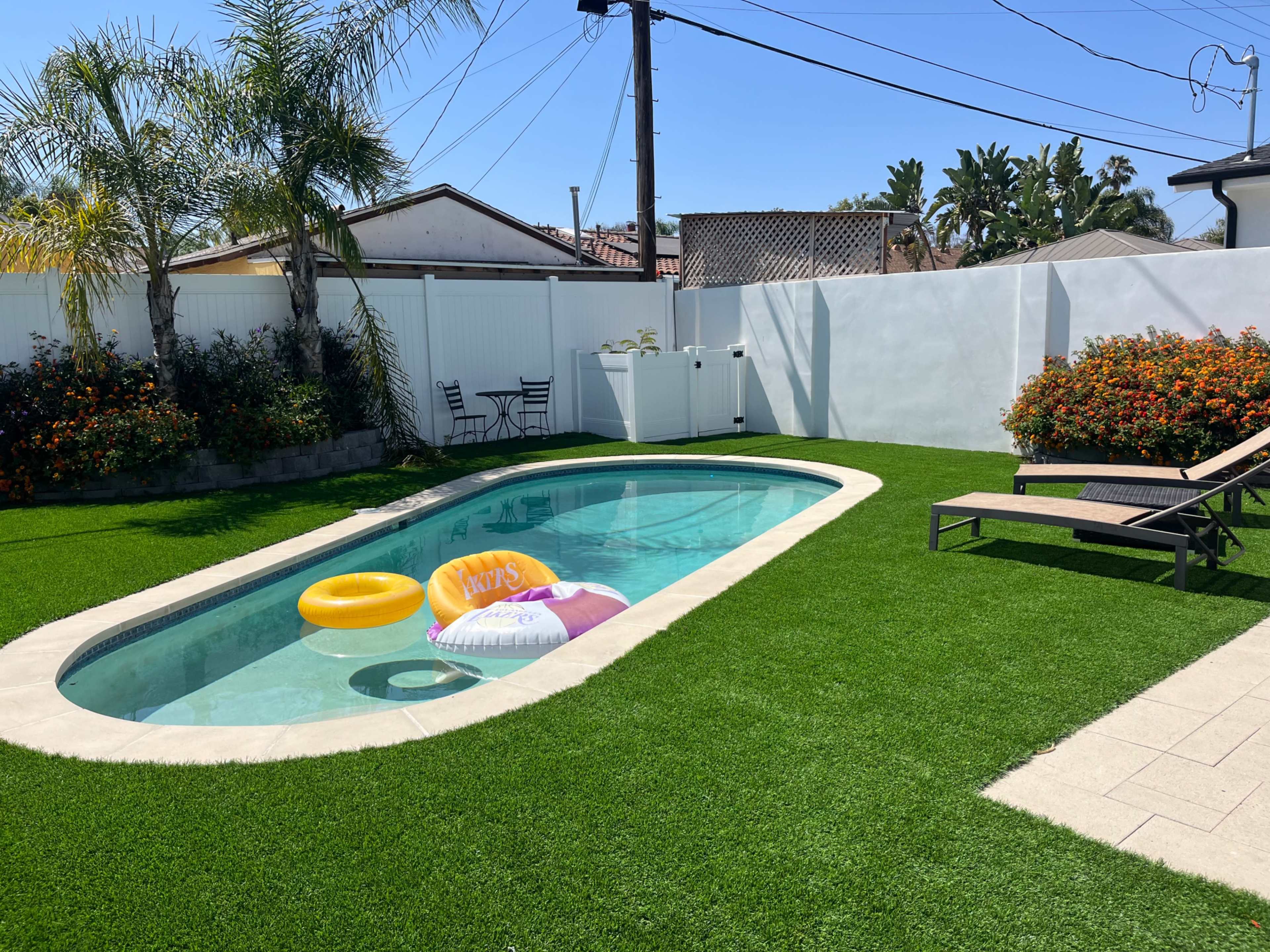 A backyard with a swimming pool, a yellow inflatable ring, lounge chairs, and vibrant flowers surrounded by artificial grass.