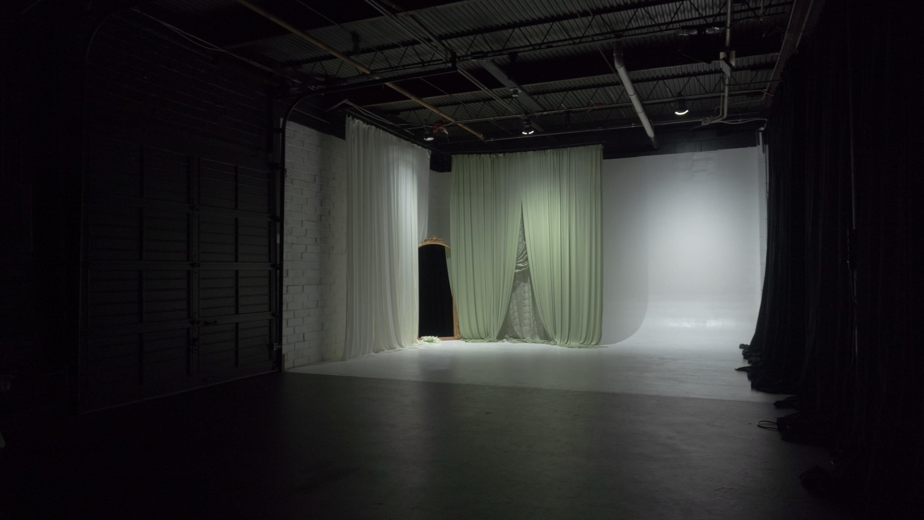 A dimly lit studio space features soft green curtains on one side and a blank white backdrop on the other.