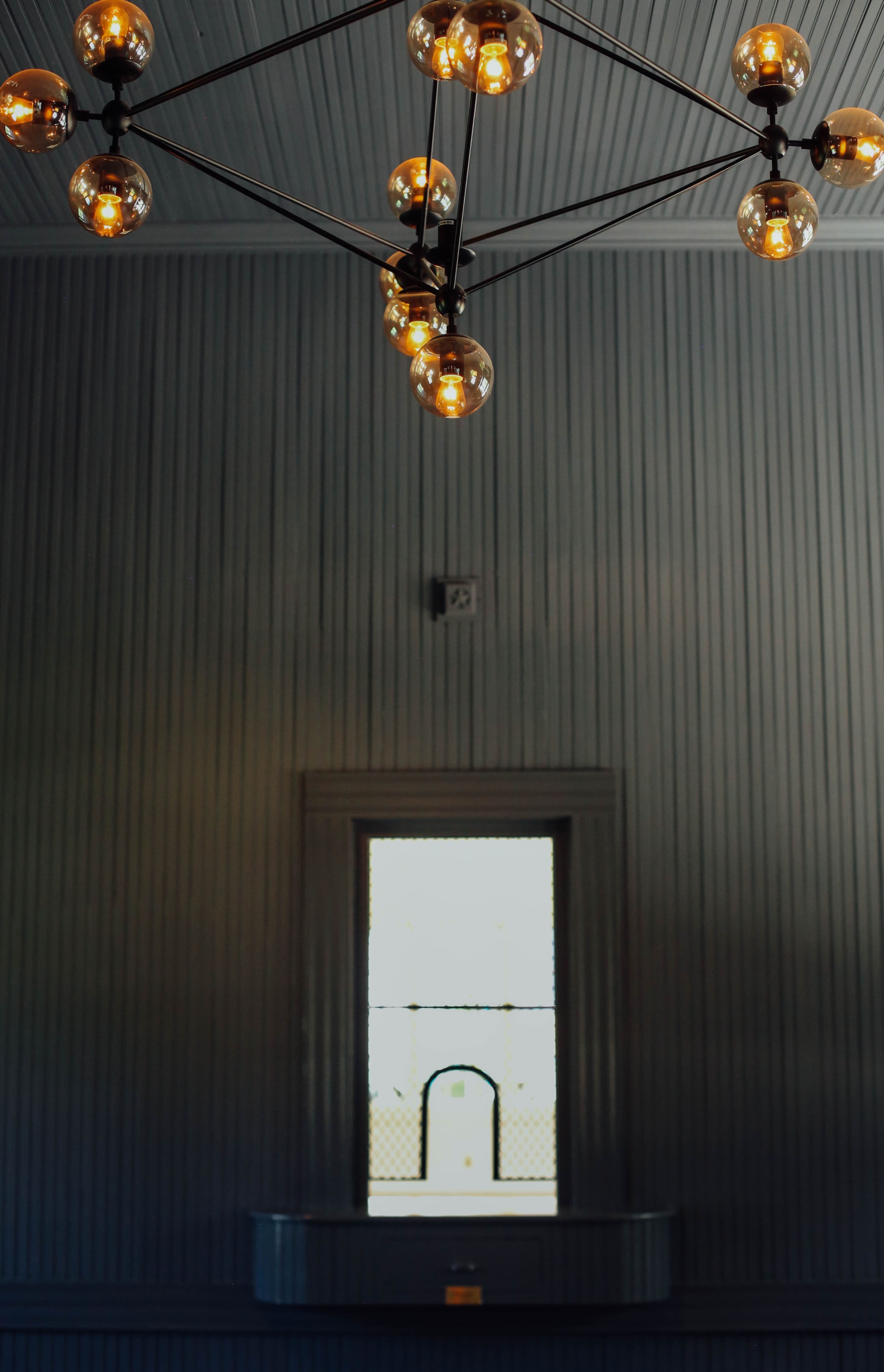 A chandelier with spherical light bulbs hangs from a ceiling above a window with a grid pattern, situated in a room with striped walls.