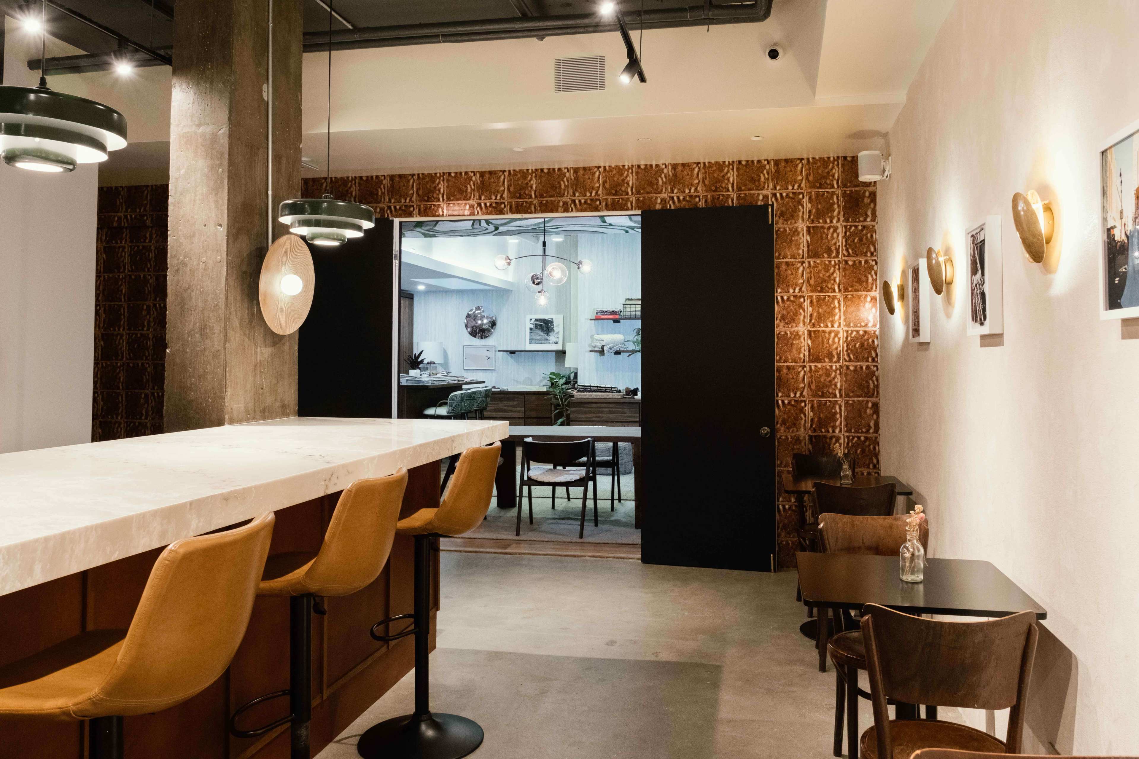 A modern café interior with a marble counter, wooden bar stools, and a view into a bright room with framed artwork and plants.
