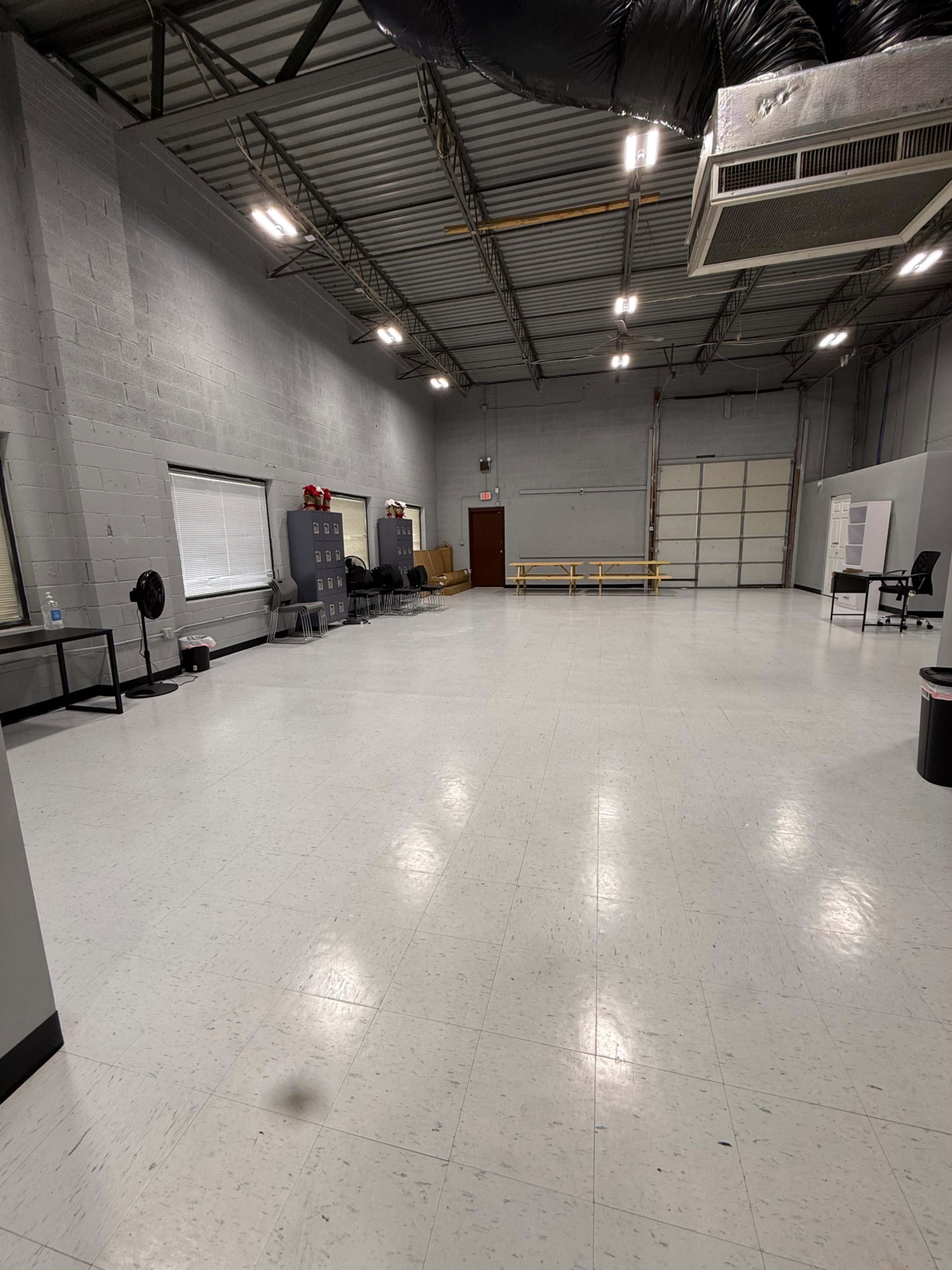 The image shows a large, empty room with gray walls, a smooth floor, a single fan, and a few pieces of furniture along one side.