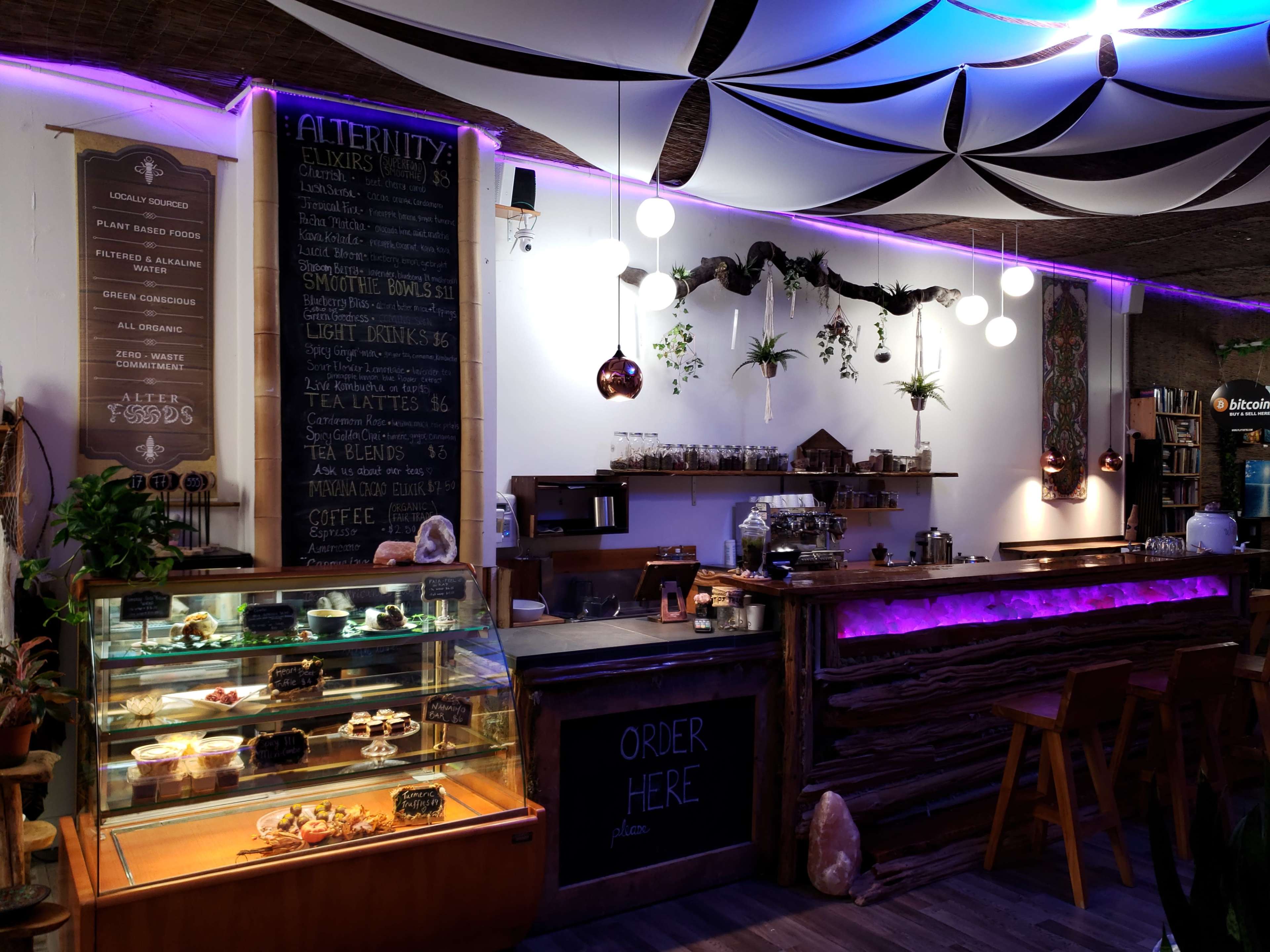 The image shows a cozy café interior featuring a counter with a display case of desserts, a chalkboard menu, and decorative plants.