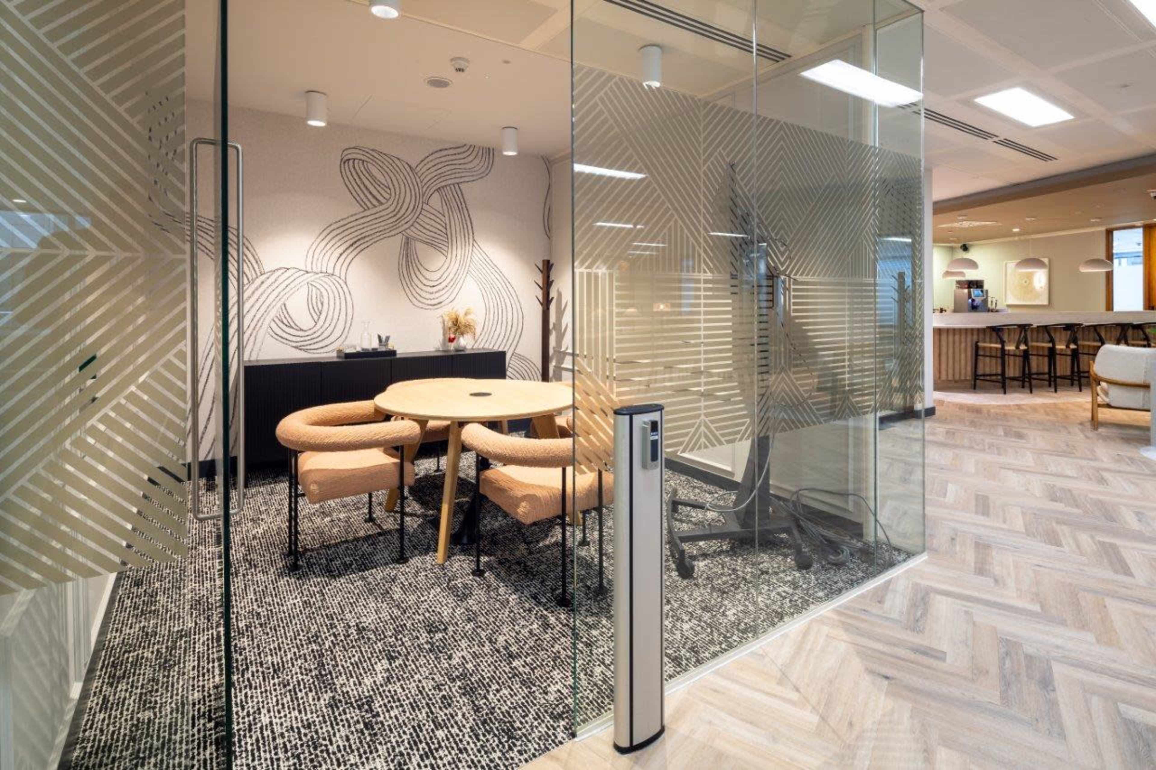 A modern office space with a glass-walled meeting room featuring a round table and stylish chairs, complemented by patterned walls and wooden flooring.