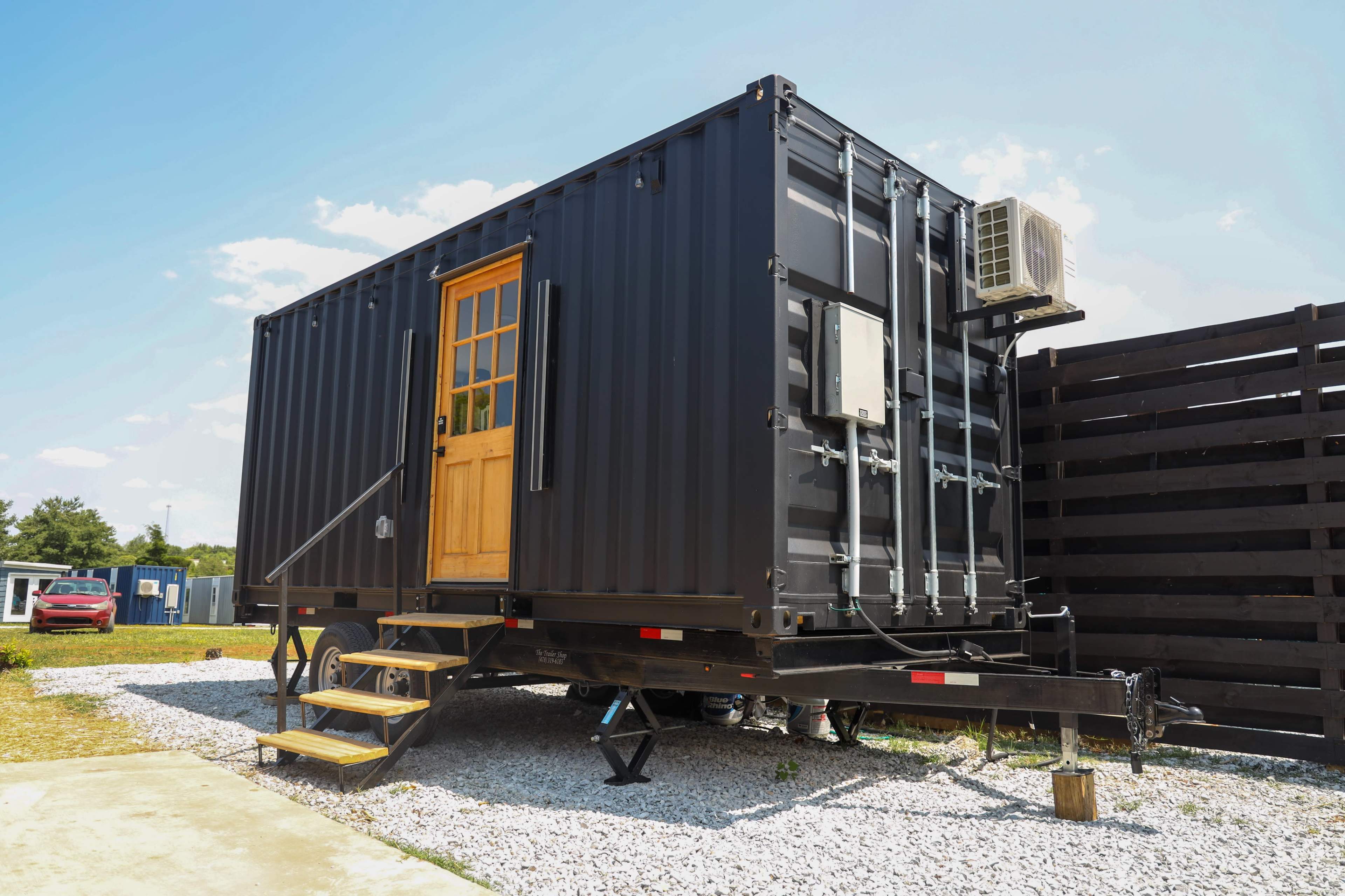 A black shipping container is set on a trailer with a wooden door and air conditioning unit, surrounded by gravel and a wooden fence.