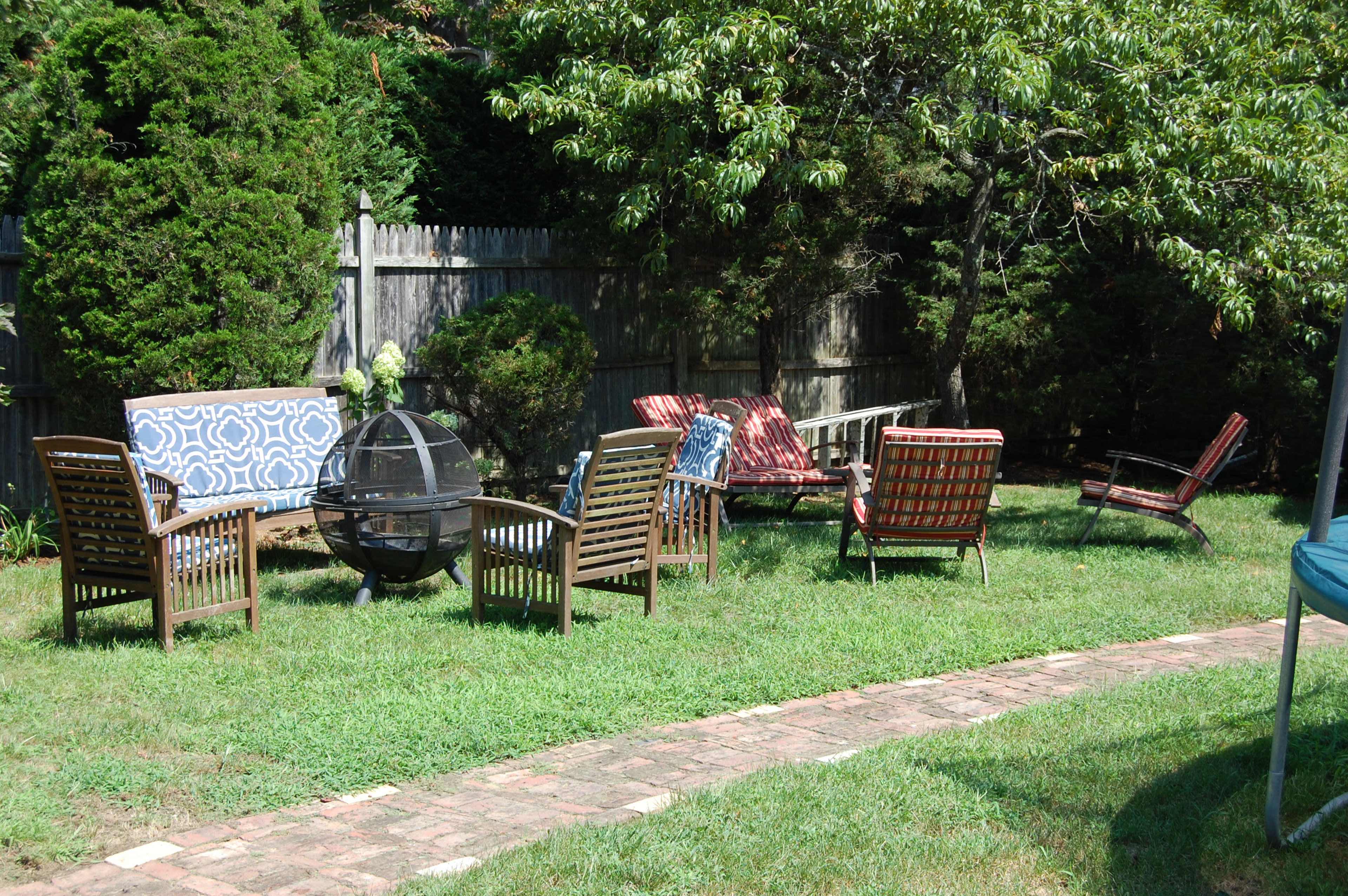 A backyard seating arrangement with wooden chairs and a circular fire pit surrounded by grass and trees.