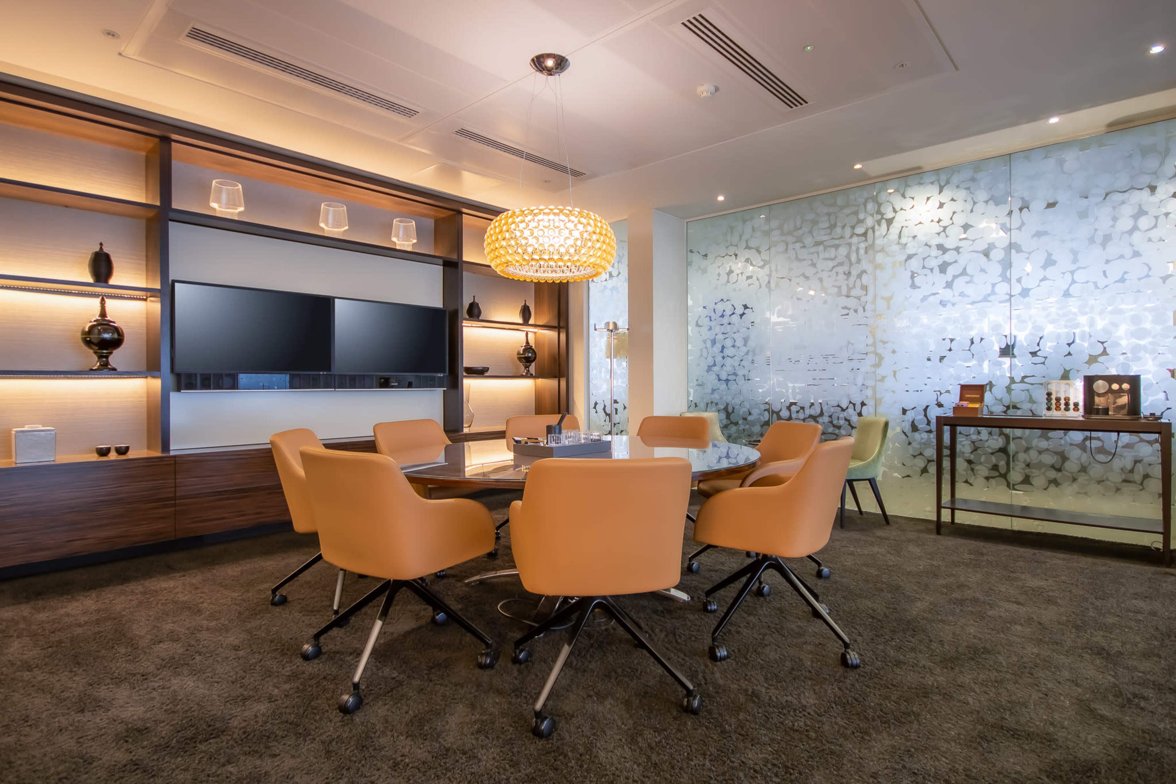 The image shows a modern meeting room featuring a round table surrounded by orange chairs, a large wall-mounted screen, and decorative lighting.