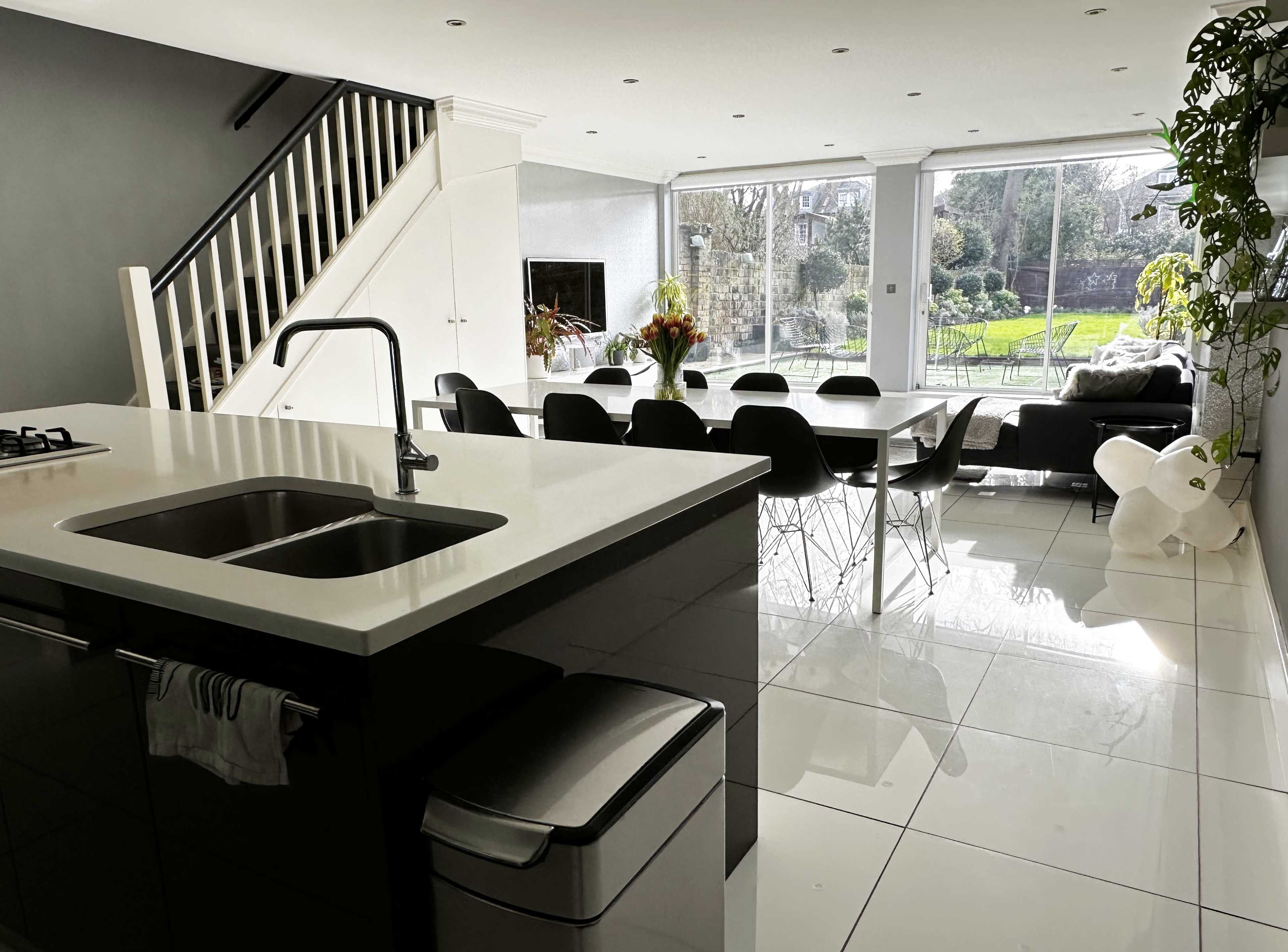 A modern, open-concept kitchen and dining area features a sleek island with a sink, surrounded by black chairs and large windows overlooking a garden.