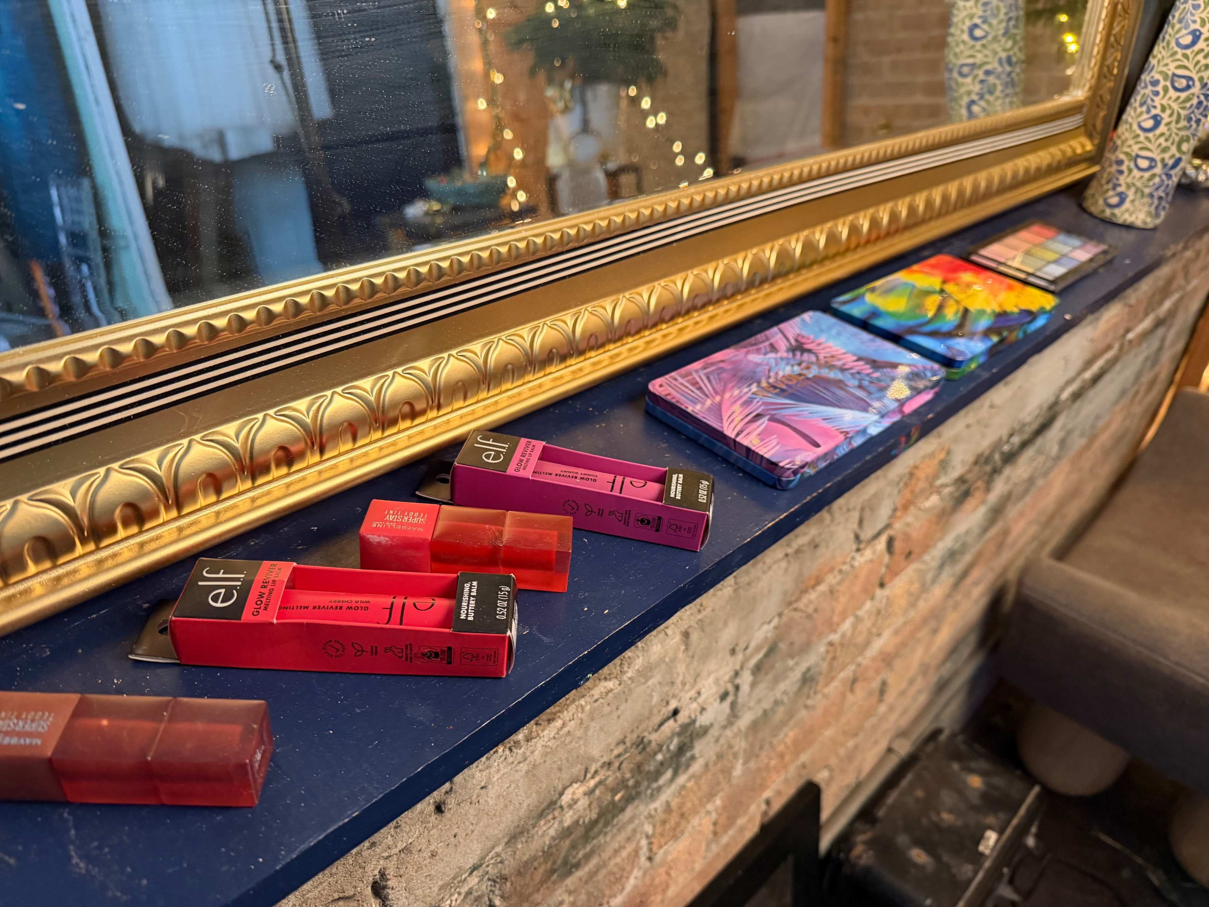The image shows a makeup display on a blue shelf featuring various ELF lipsticks and colorful palettes, with a golden framed mirror reflecting the background.