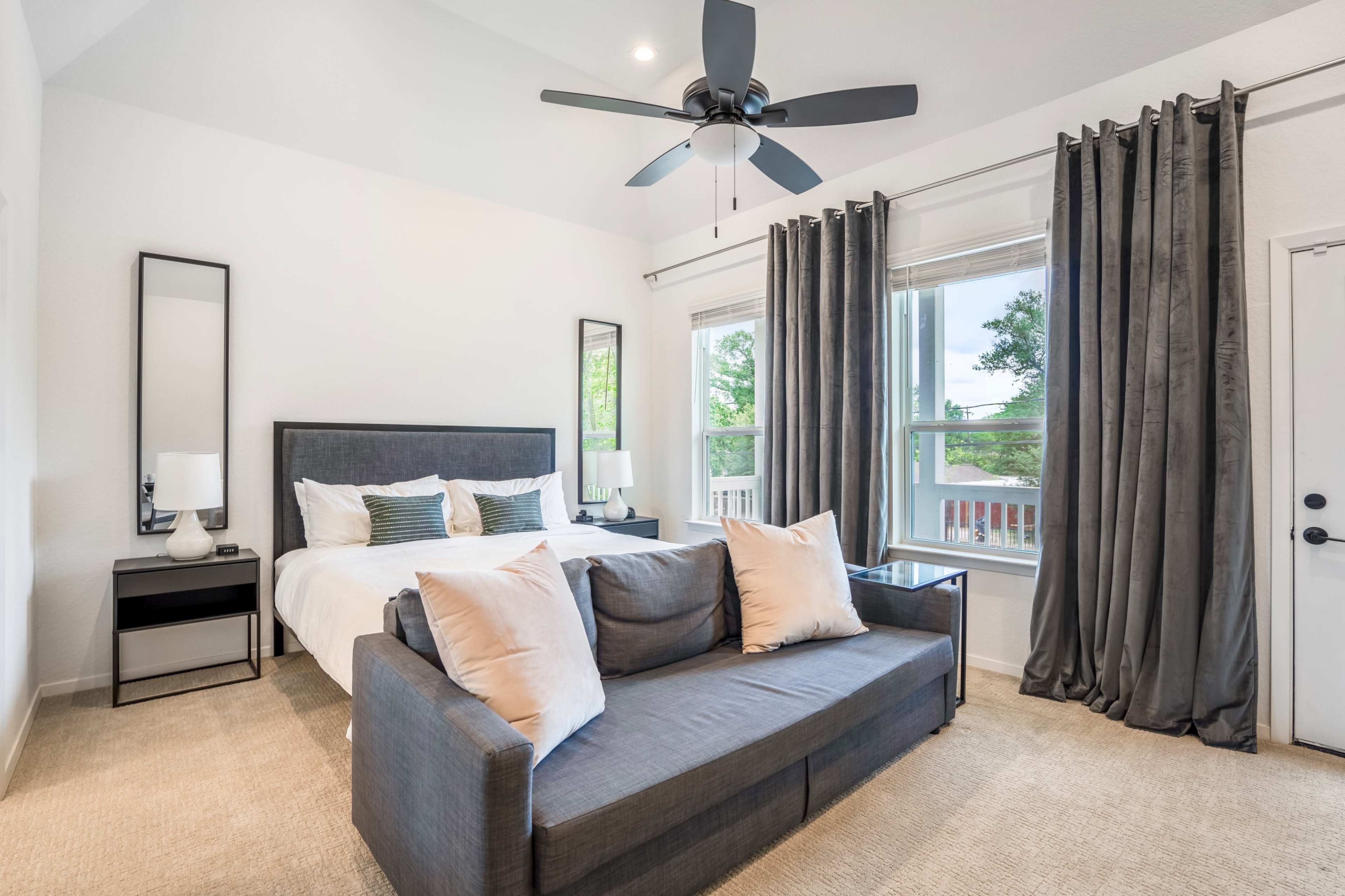 A well-lit bedroom with a king-size bed, a gray sofa with pillows, and large windows covered by curtains.