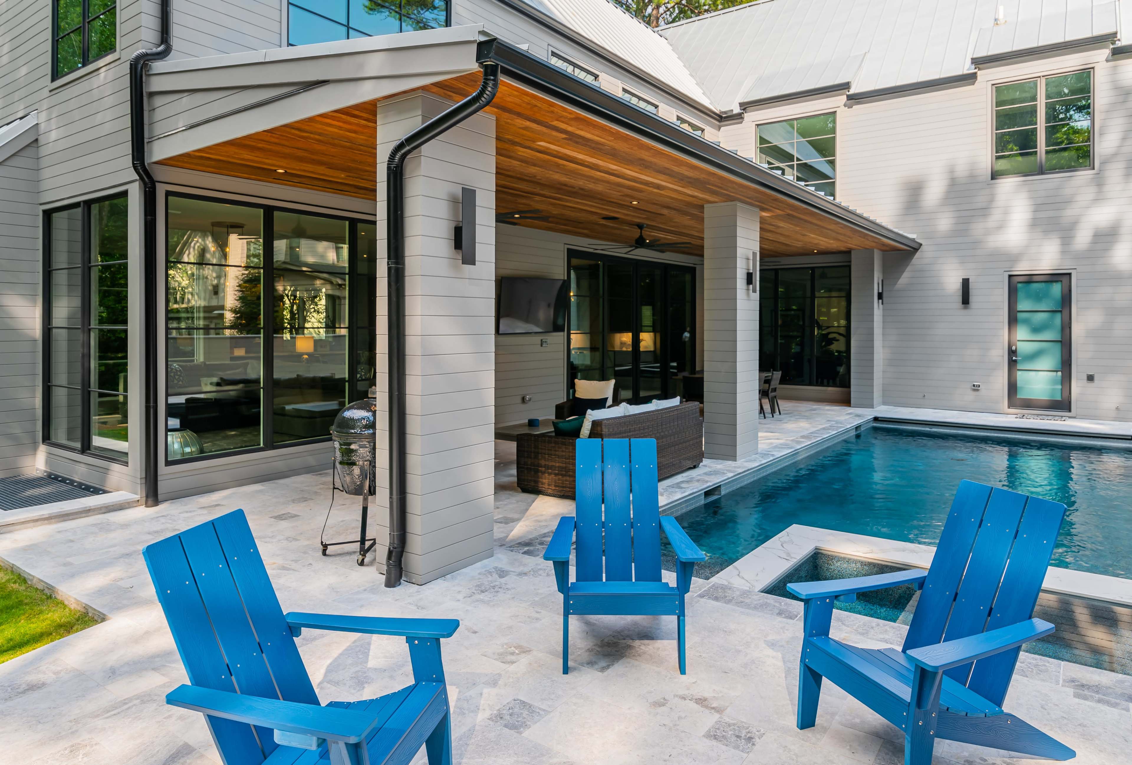 The image shows a modern house with an outdoor seating area near a swimming pool, featuring blue adirondack chairs arranged in front of a covered patio with large glass windows.