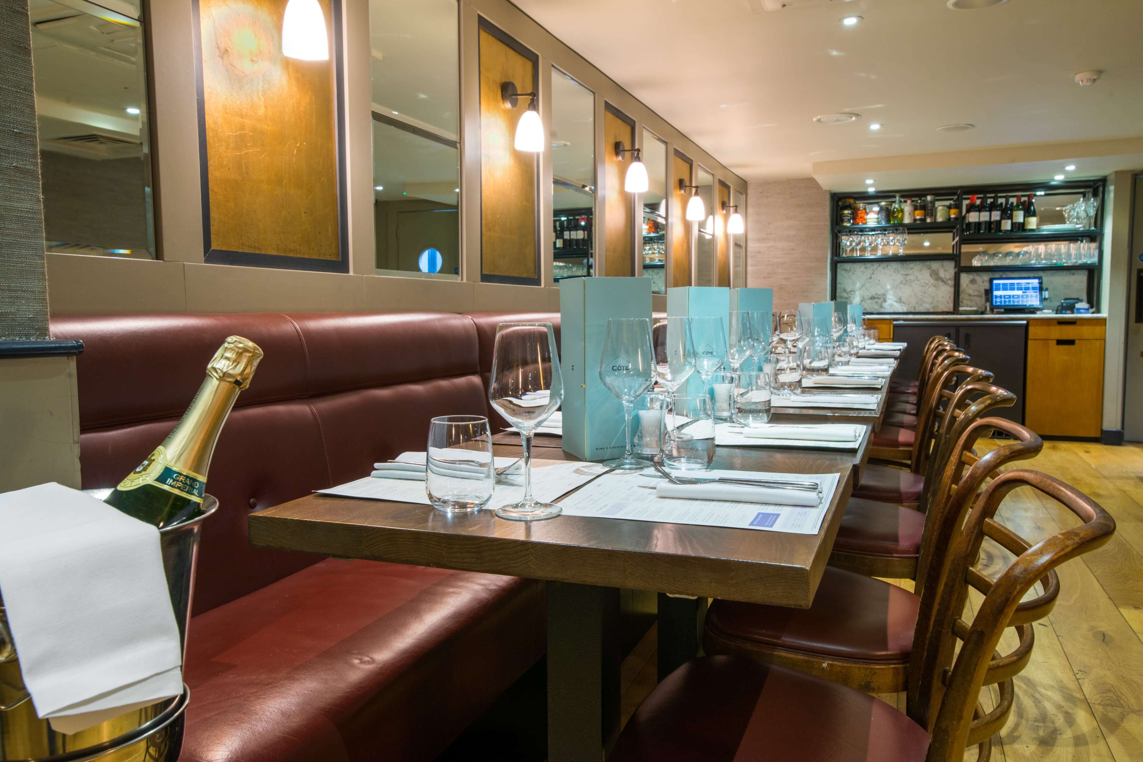 A neatly arranged dining table with wine glasses, menus, and a bottle of champagne on ice is set in a modern restaurant interior.